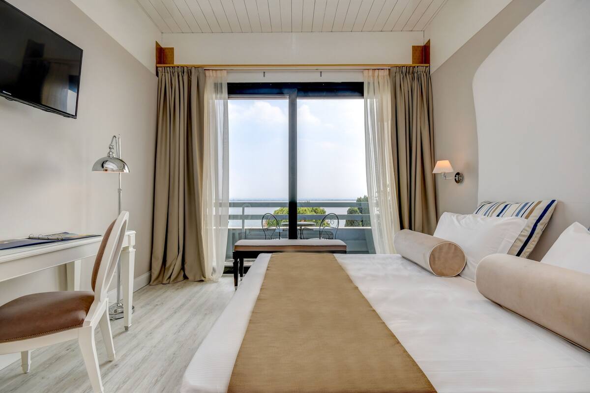 A hotel room with a bed, a desk, a chair, a TV, and a balcony with a view.