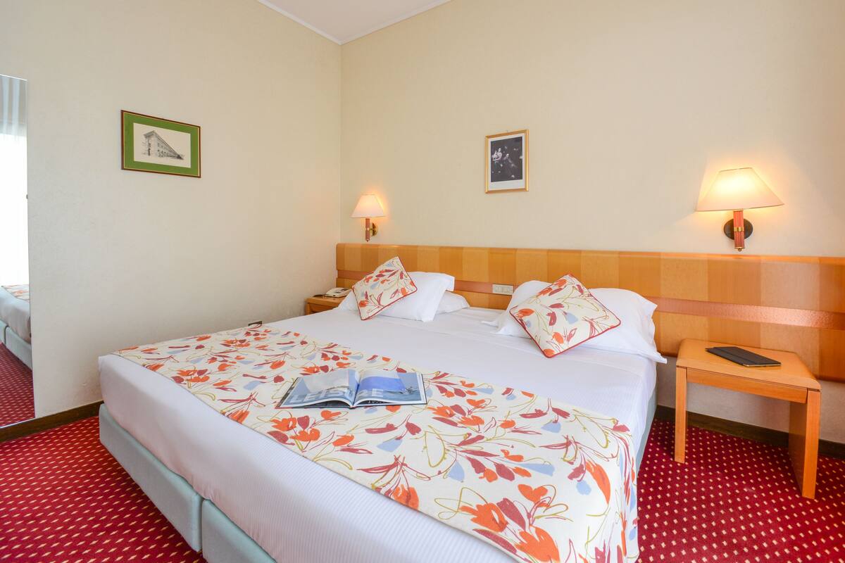A hotel room with a large bed, two bedside tables, a nightstand, a red carpeted floor, and two wall-mounted lamps.