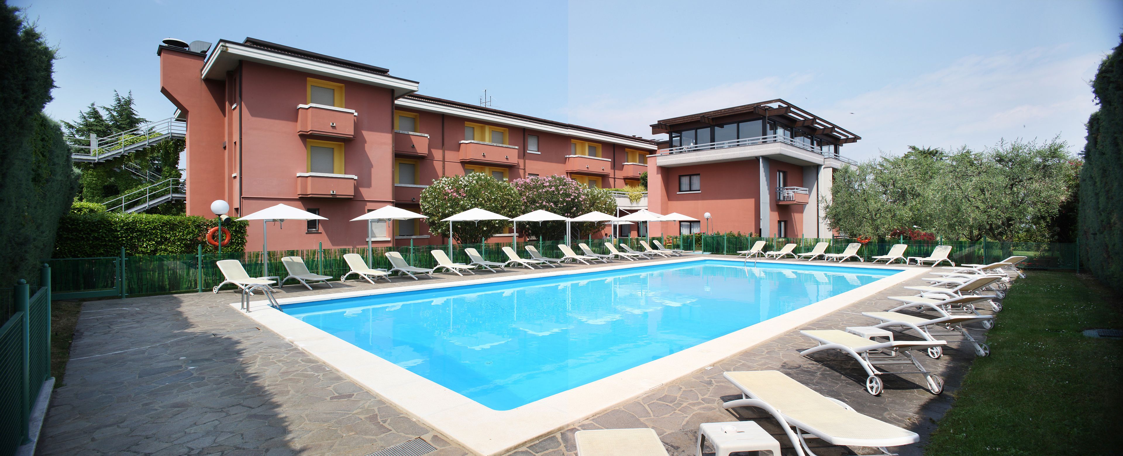 Una piscina con tumbonas y sombrillas, rodeada por un edificio con balcones y un jardín.