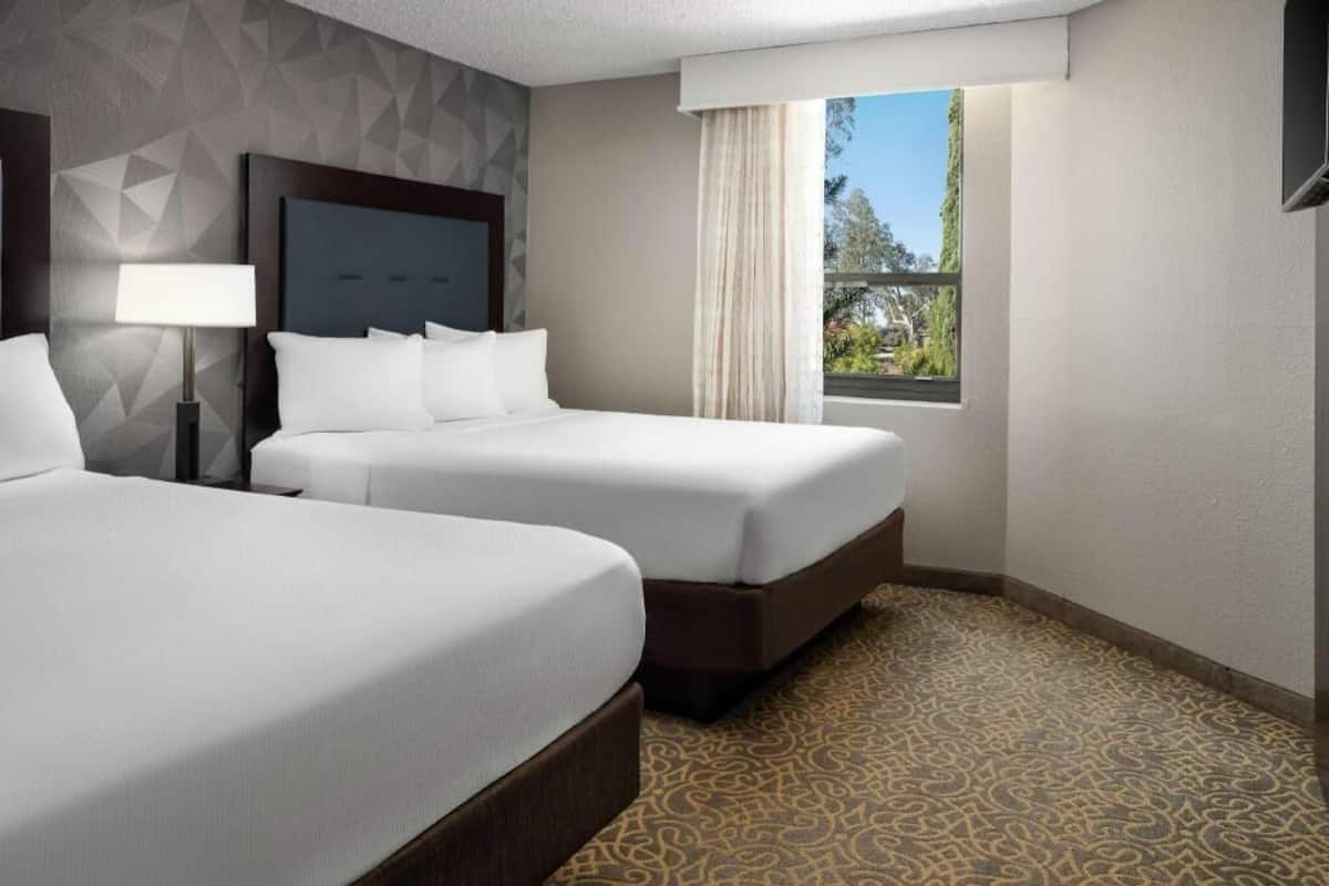 A hotel room with two beds, a patterned carpet, a window with a view, and a geometric wall design.