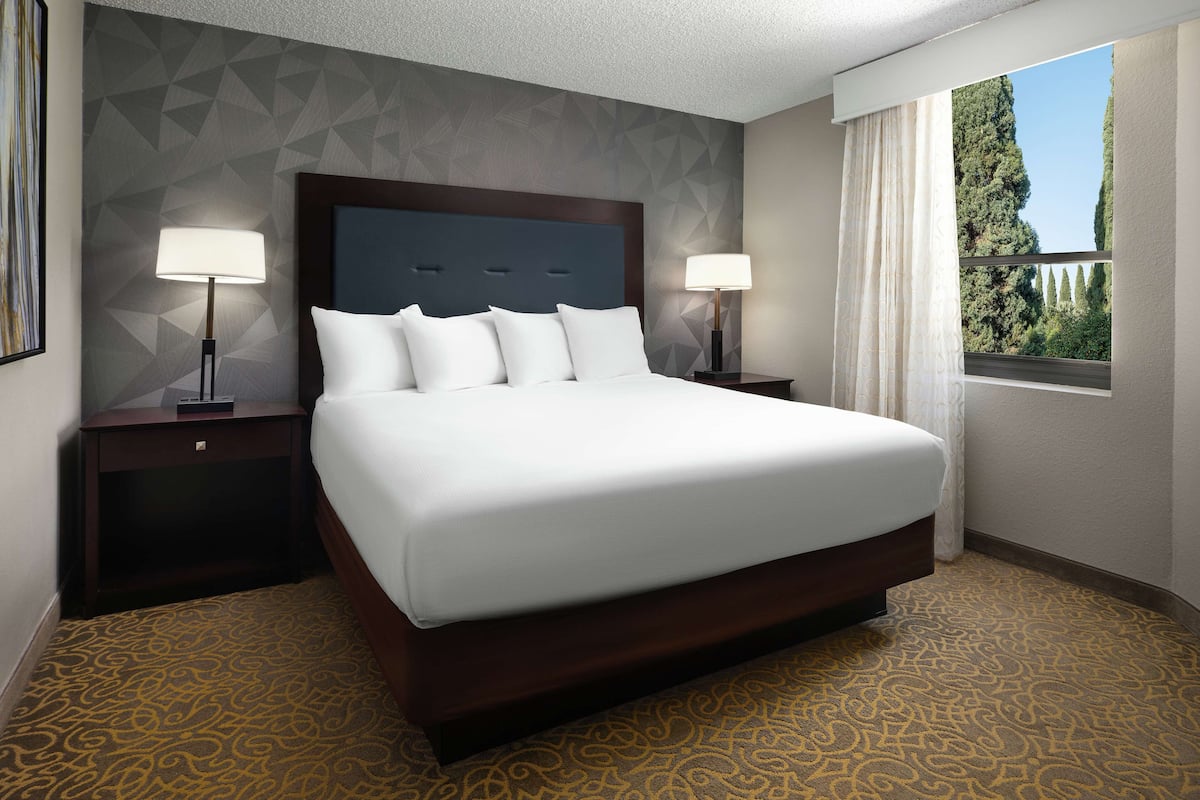 A hotel room with a large bed, two bedside tables, a headboard, and a window with curtains.