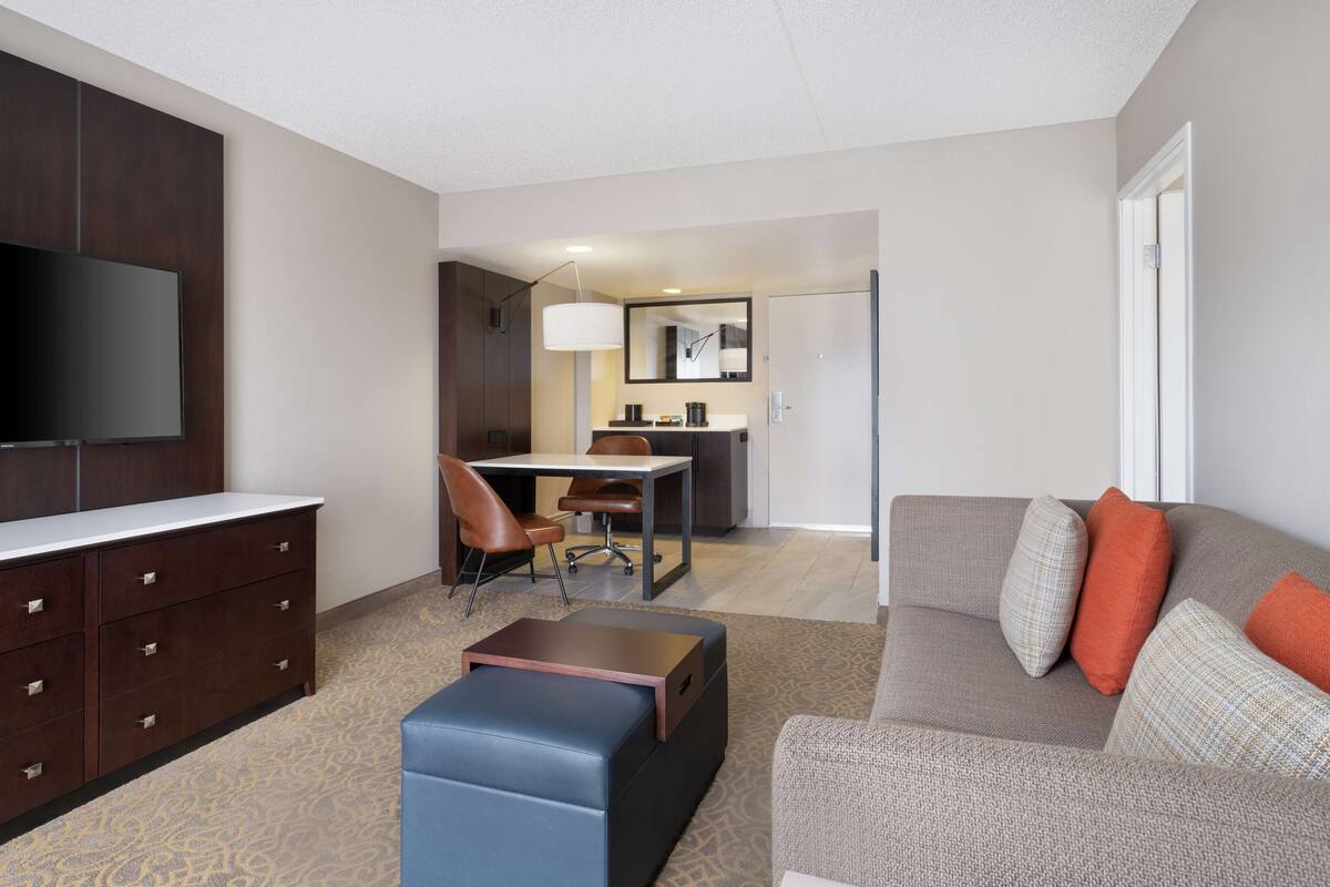 A modern living room with a flat-screen TV, a sofa with orange cushions, a coffee table, and a desk with a chair.