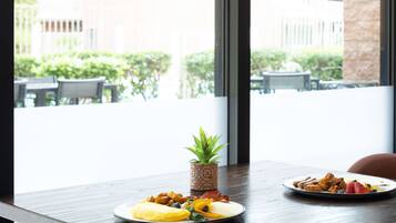 A dining table with plates of food and a small plant, set against a window with a view of buildings and greenery outside.