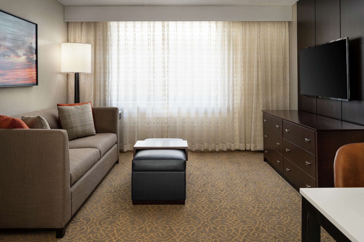 A hotel room with a sofa, ottoman, TV, and a desk.
