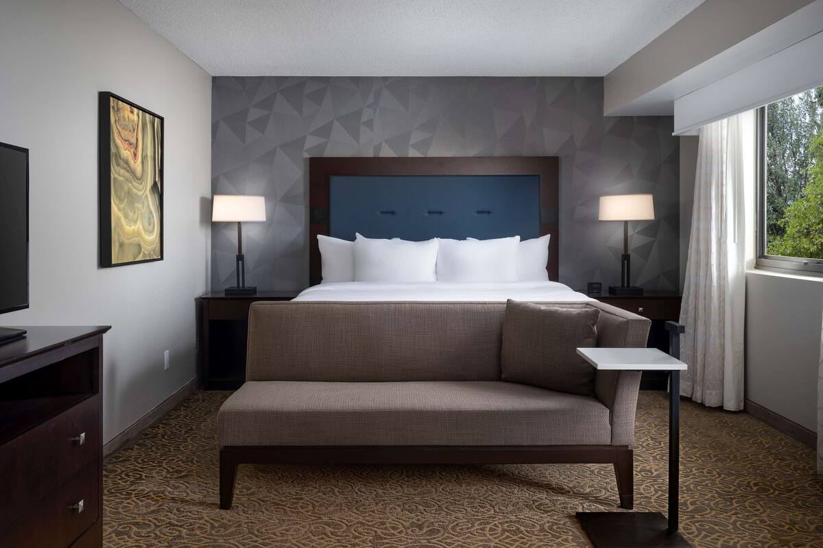 A hotel room with a large bed, a bench, two bedside tables with lamps, a TV, and a patterned carpet.