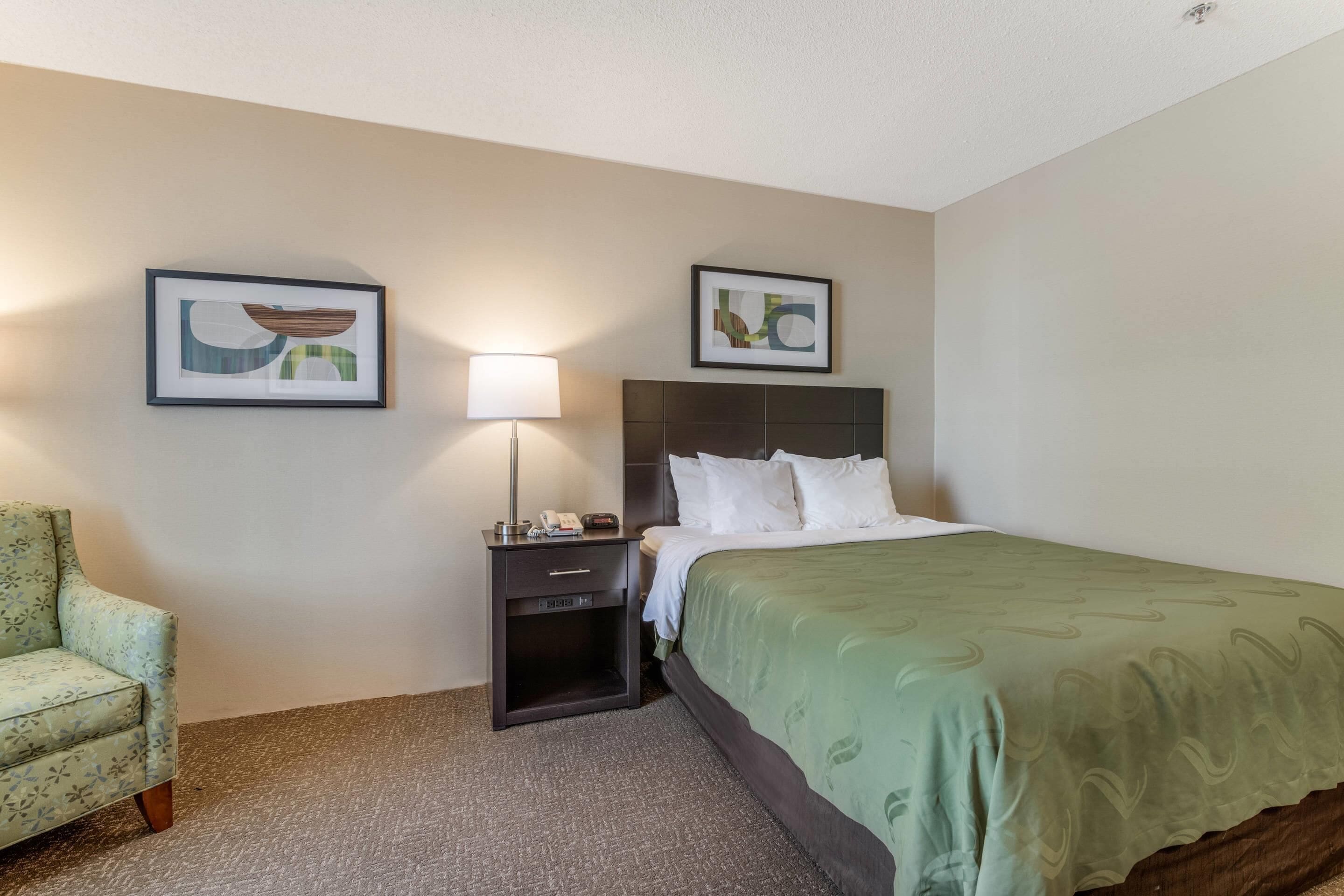A hotel room with a bed, a nightstand, a lamp, a chair, and two framed pictures on the wall.