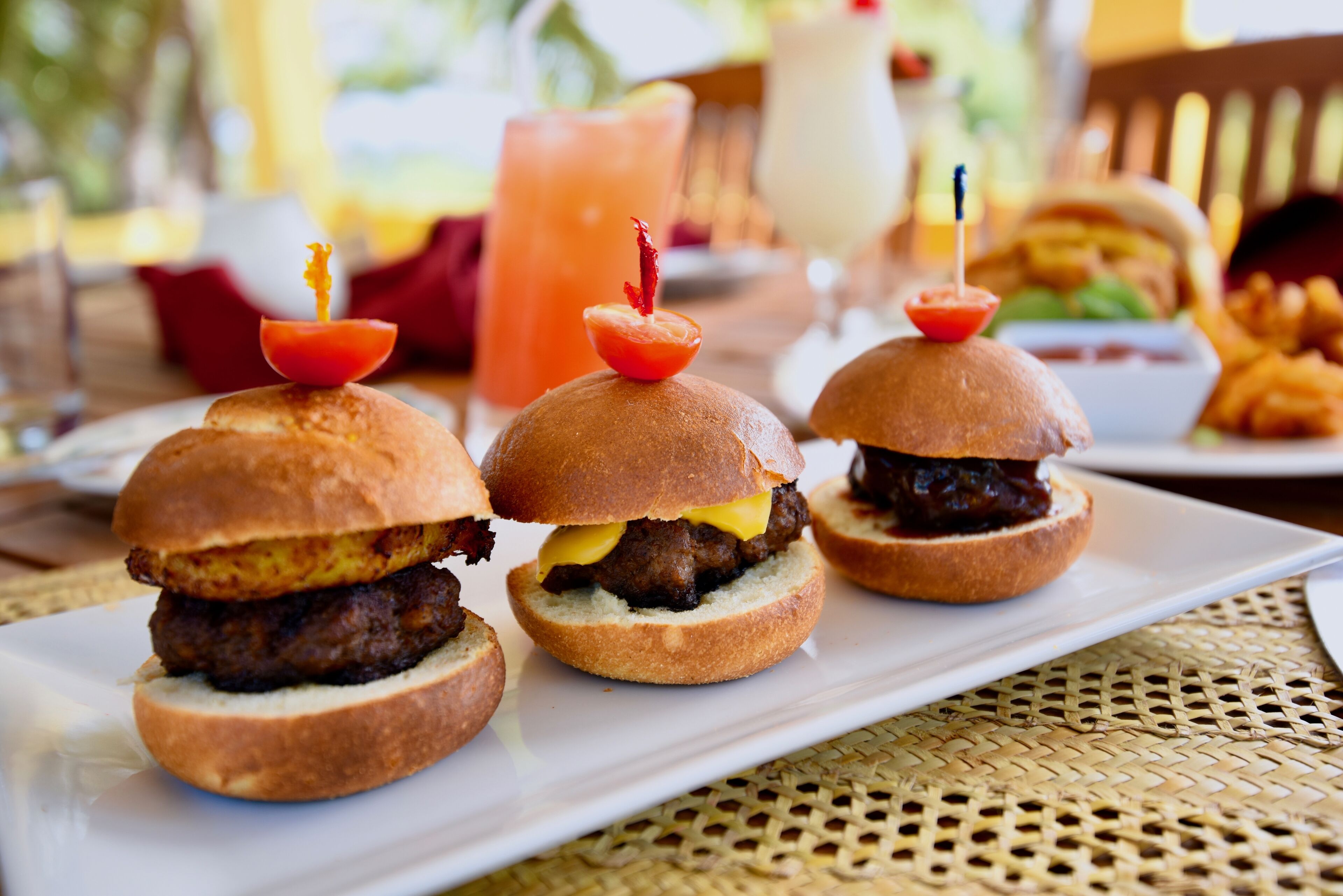 Three mini burgers with toothpicks on a white plate, a drink, and a burger in the background.