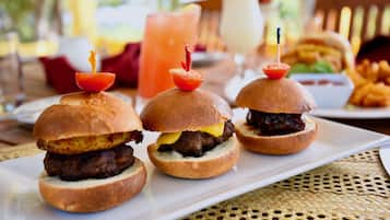 Three mini burgers with toothpicks on a white plate, a drink, and a burger in the background.