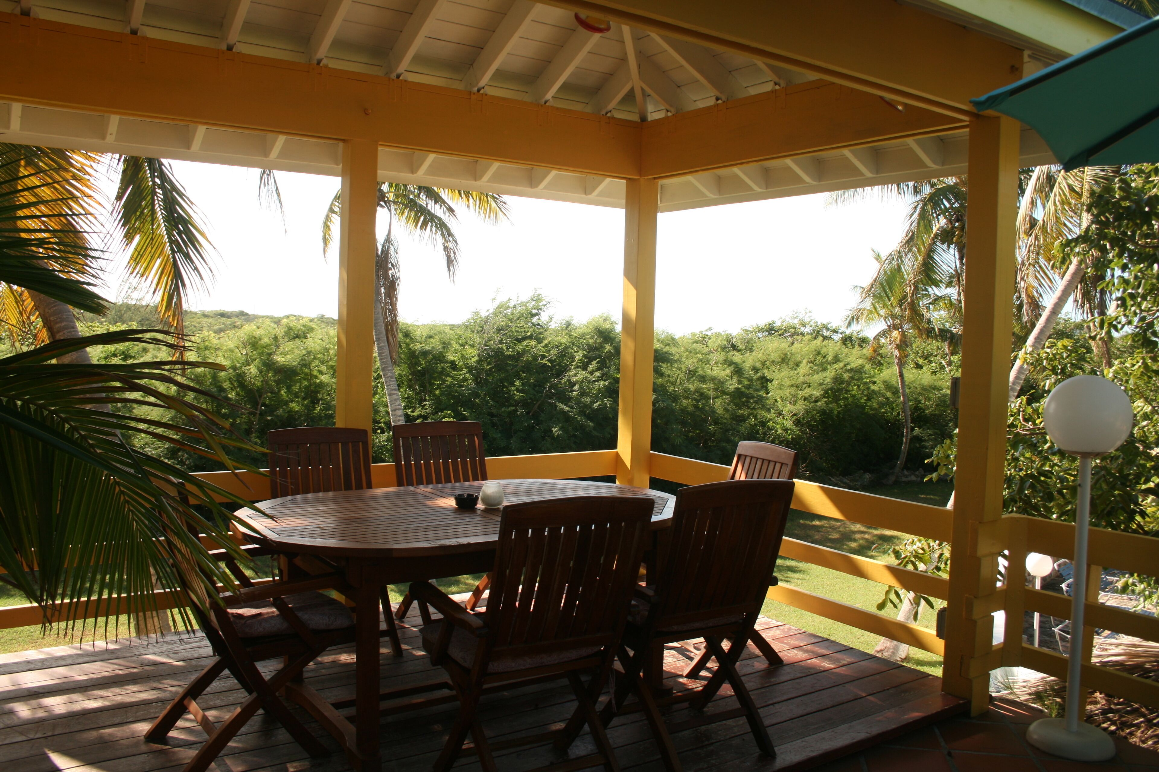 A covered outdoor seating area with a wooden table and chairs, surrounded by a yellow railing, overlooking a lush green landscape.