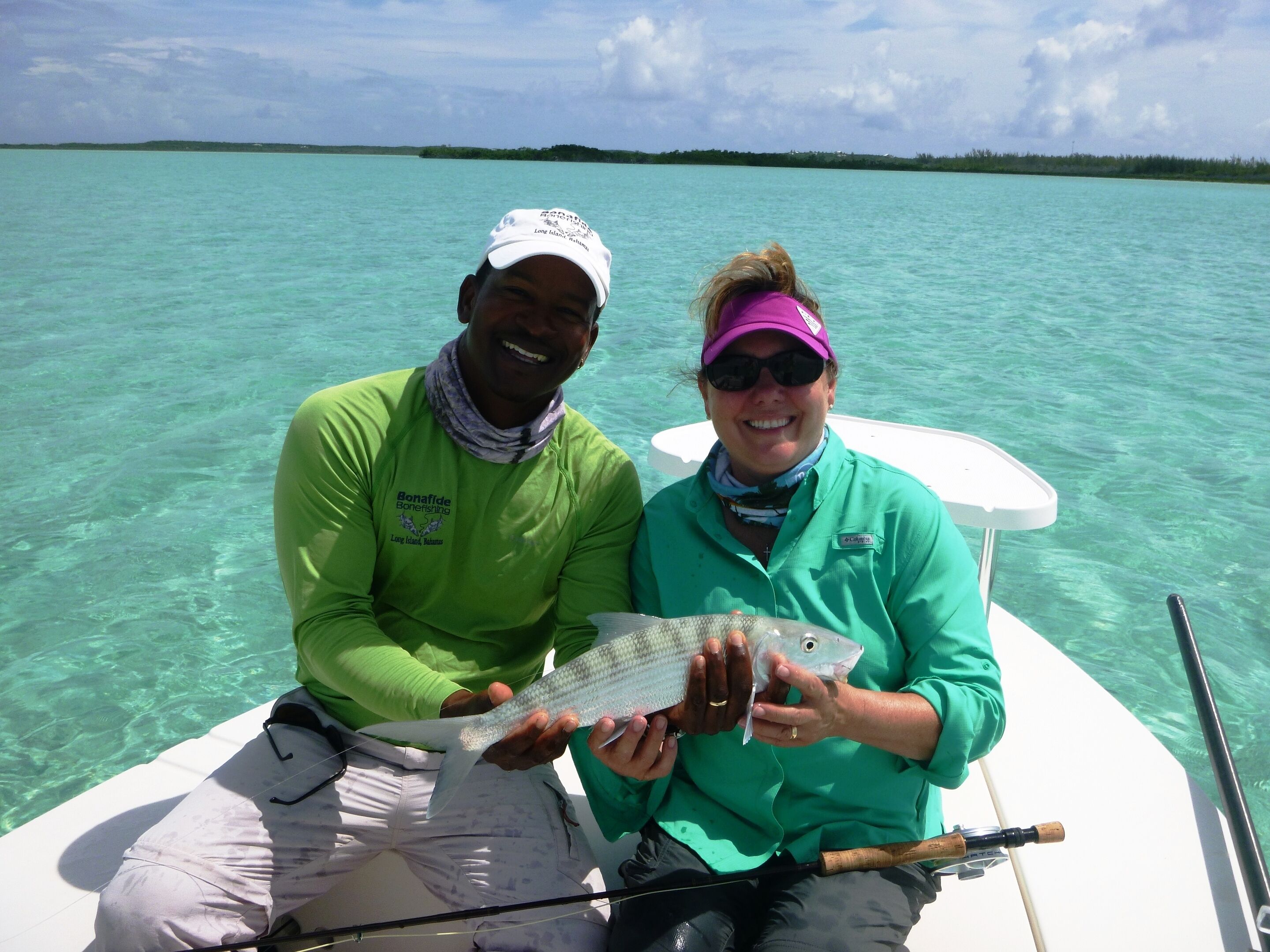 Two people on a boat holding a fish, with clear turquoise water and a distant shoreline.