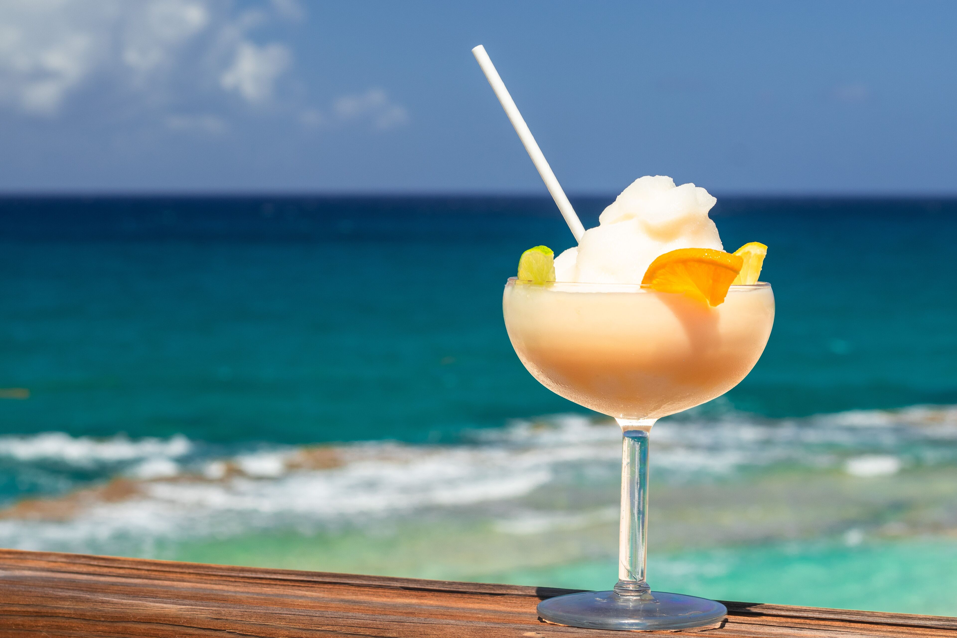Ein Cocktailglas mit Trinkhalm, garniert mit einer Limettenscheibe und einer Orangenscheibe, auf einer Holzoberfläche mit Blick auf das Meer.
