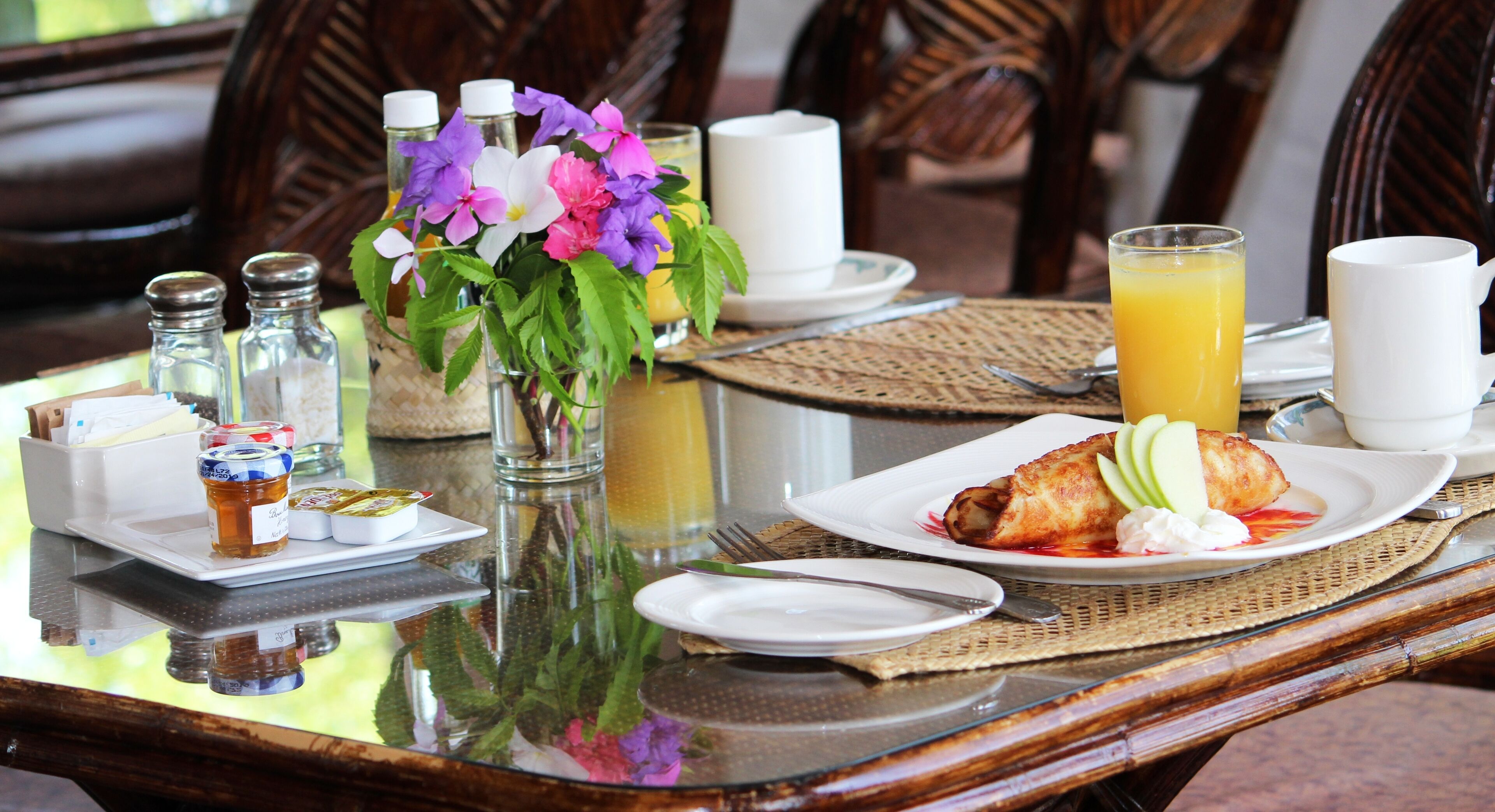 A breakfast table with a croissant, fruit, and juice. There are salt and pepper shakers, a vase with flowers, and a glass of orange juice.