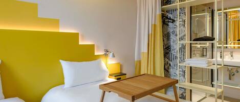 A hotel room with a bed, bedside table, and a small wooden table. There is a mirror with a patterned curtain and a shelf with towels.