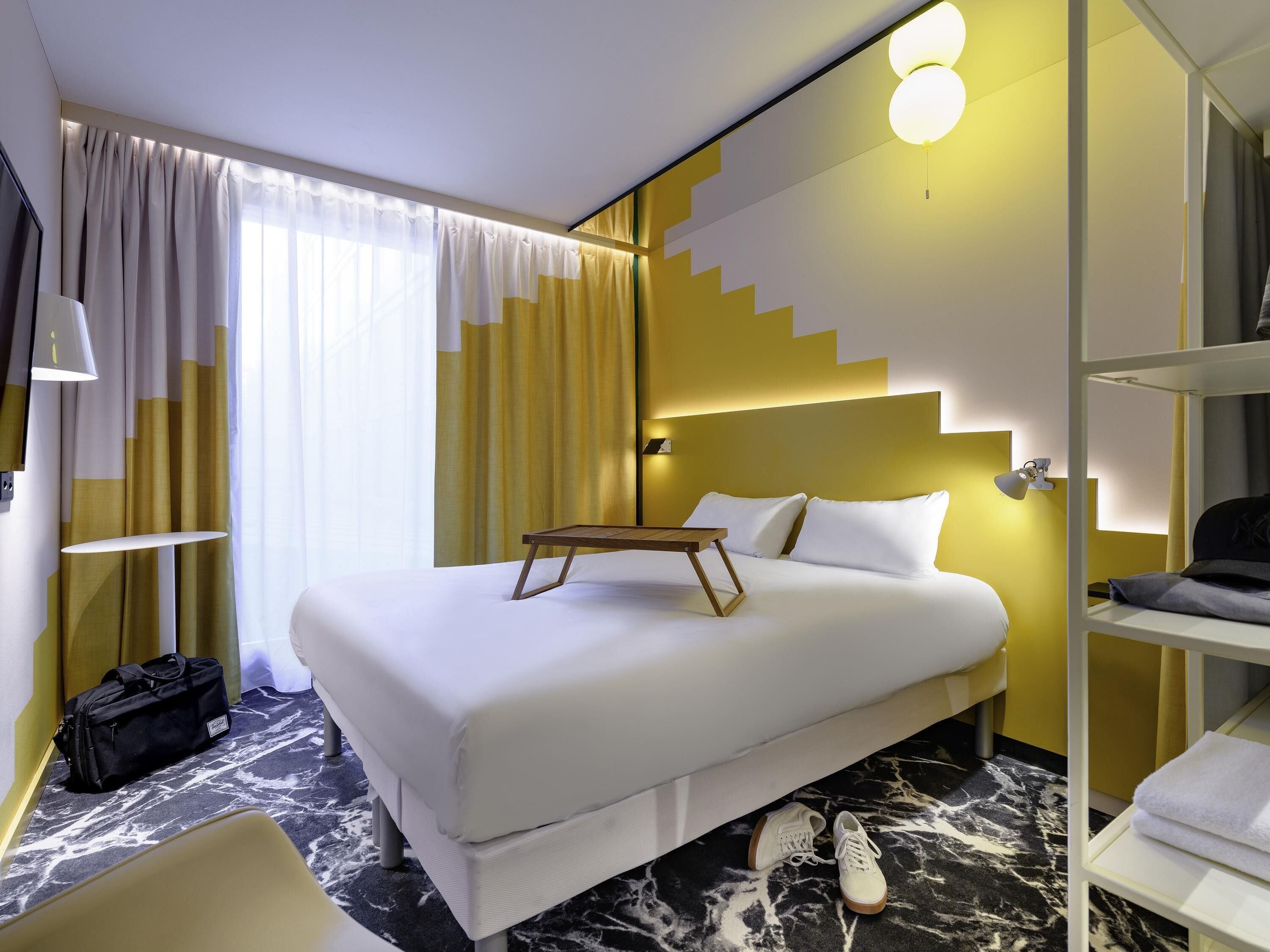 A hotel room with a bed, bedside table, lamp, suitcase, and a view of the outside.