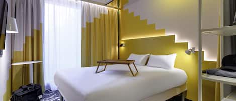 A hotel room with a bed, bedside table, lamp, suitcase, and a view of the outside.