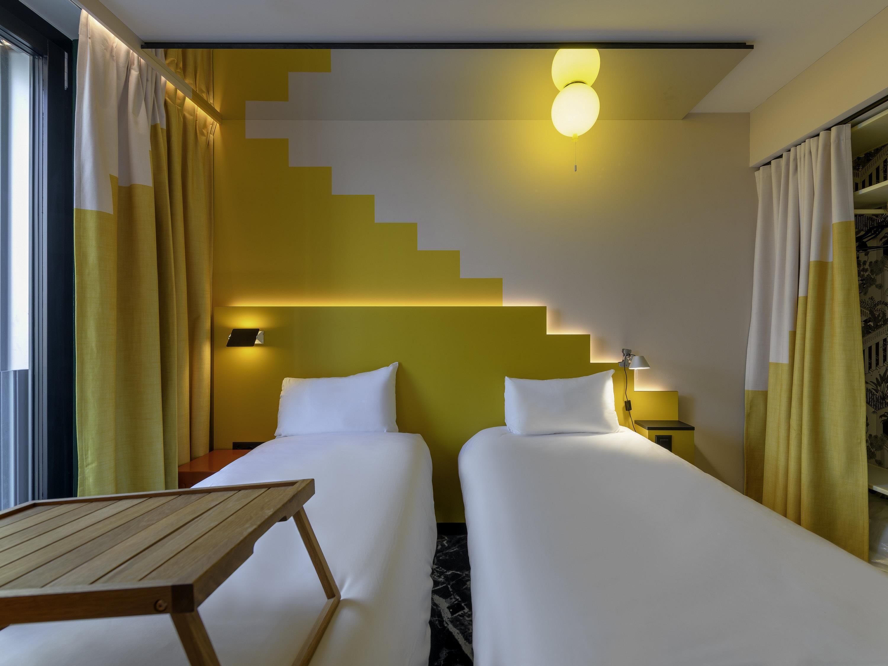 A hotel room with two beds, a wooden table, a staircase, and a ceiling light.