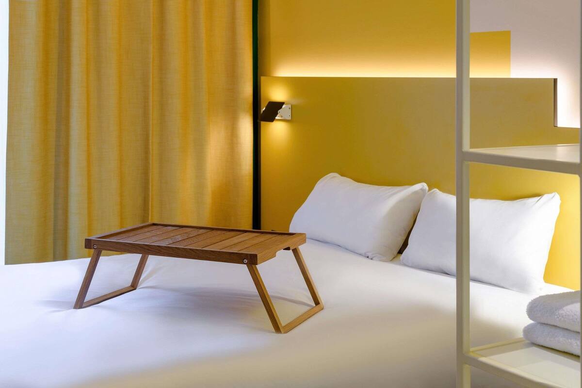 A hotel room with a bed, a wooden table, white pillows, a wall-mounted lamp, and a curtain.