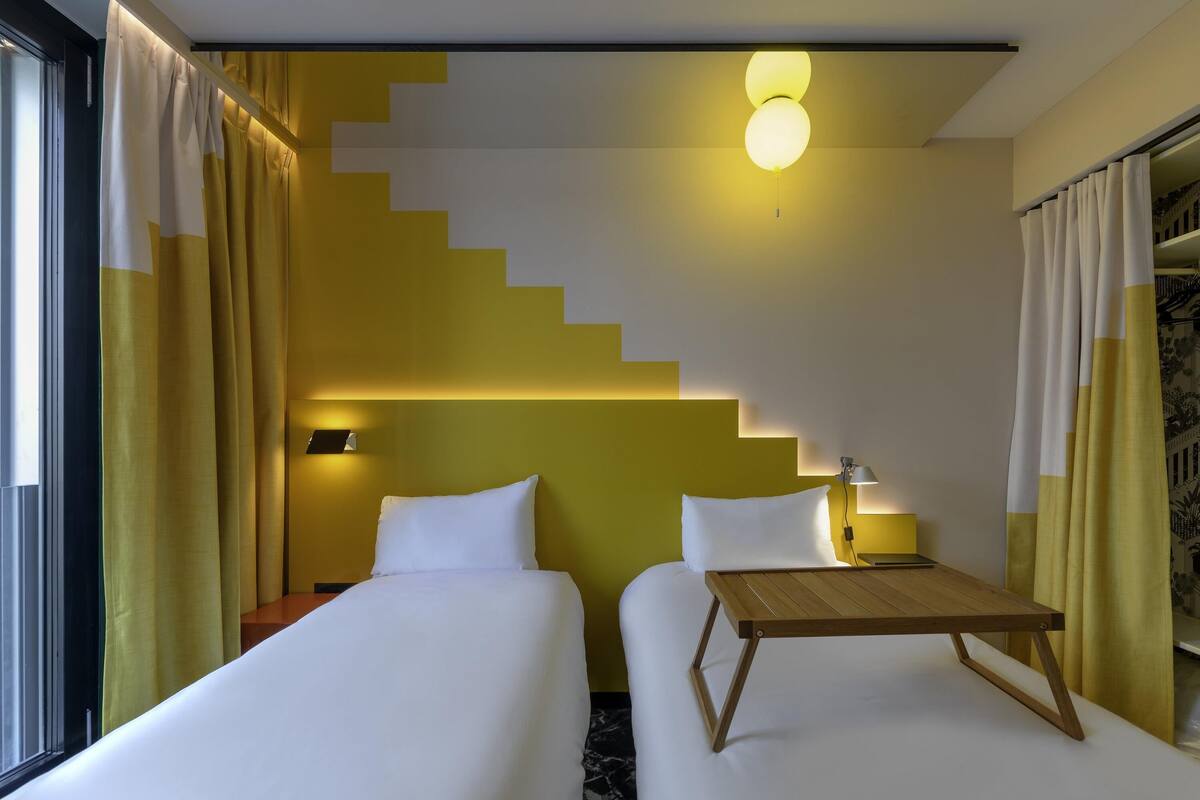 A hotel room with two beds, a staircase, a ceiling light, and a nightstand with a lamp.