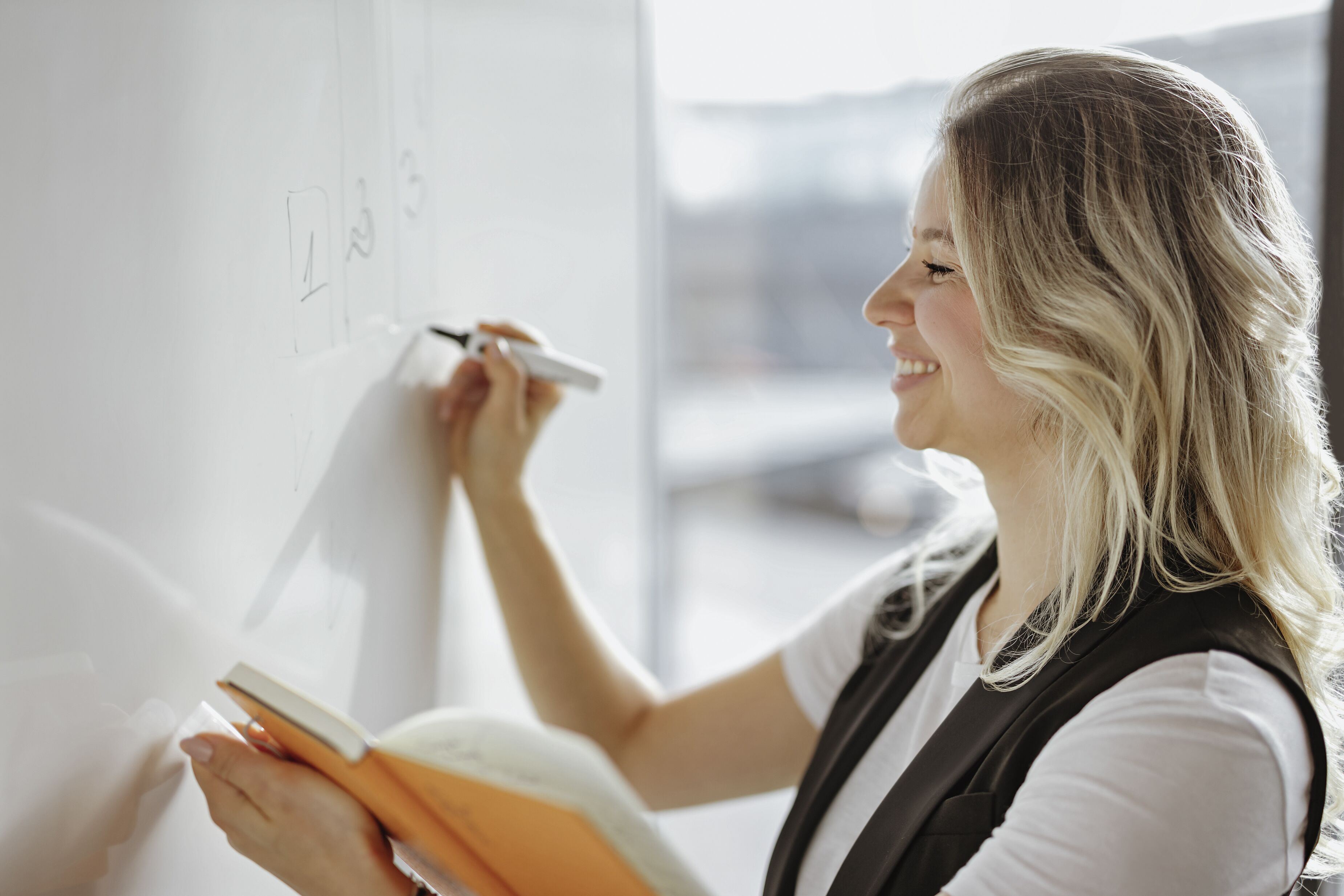 A person writing on a whiteboard with a marker, holding a book.