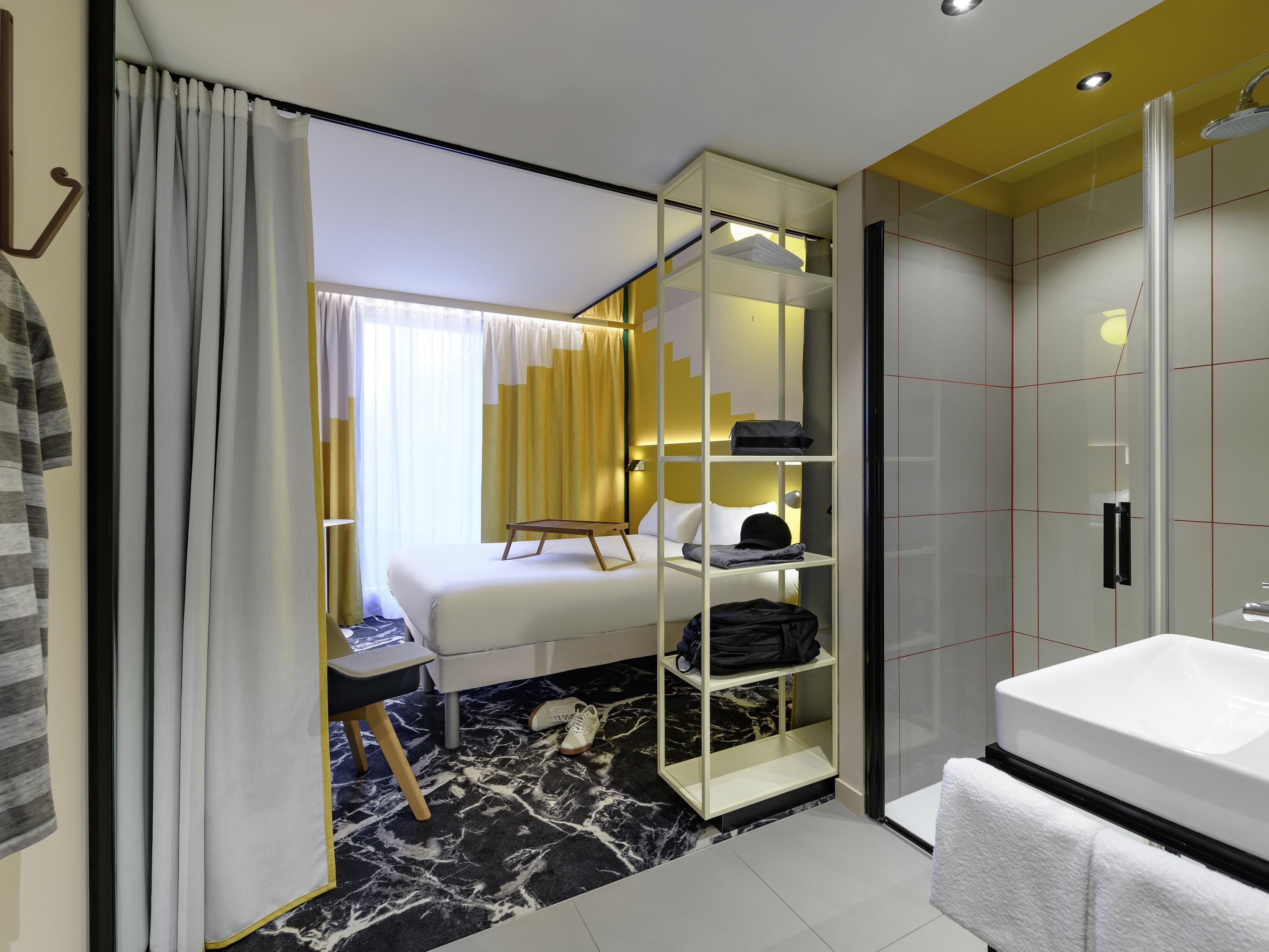 A hotel room with a bed, a white desk, a glass shelving unit, a bathroom with a white sink, and a shower.