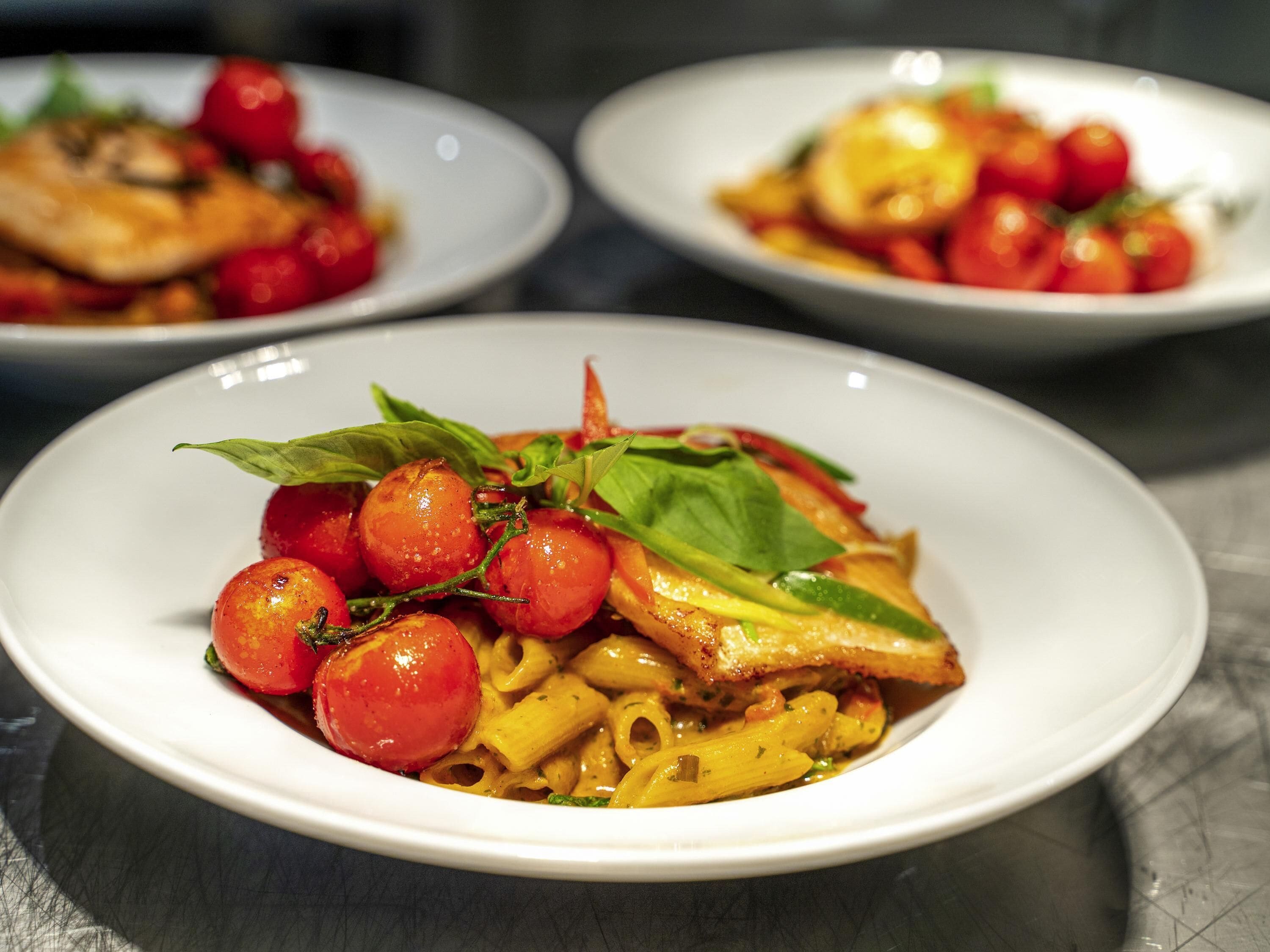 Three plates of pasta with cherry tomatoes and basil on a table.
