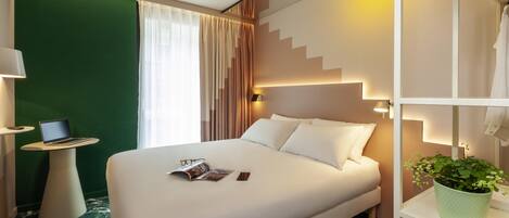 A hotel room with a bed, a small table with a laptop, a magazine, a potted plant, and a staircase.