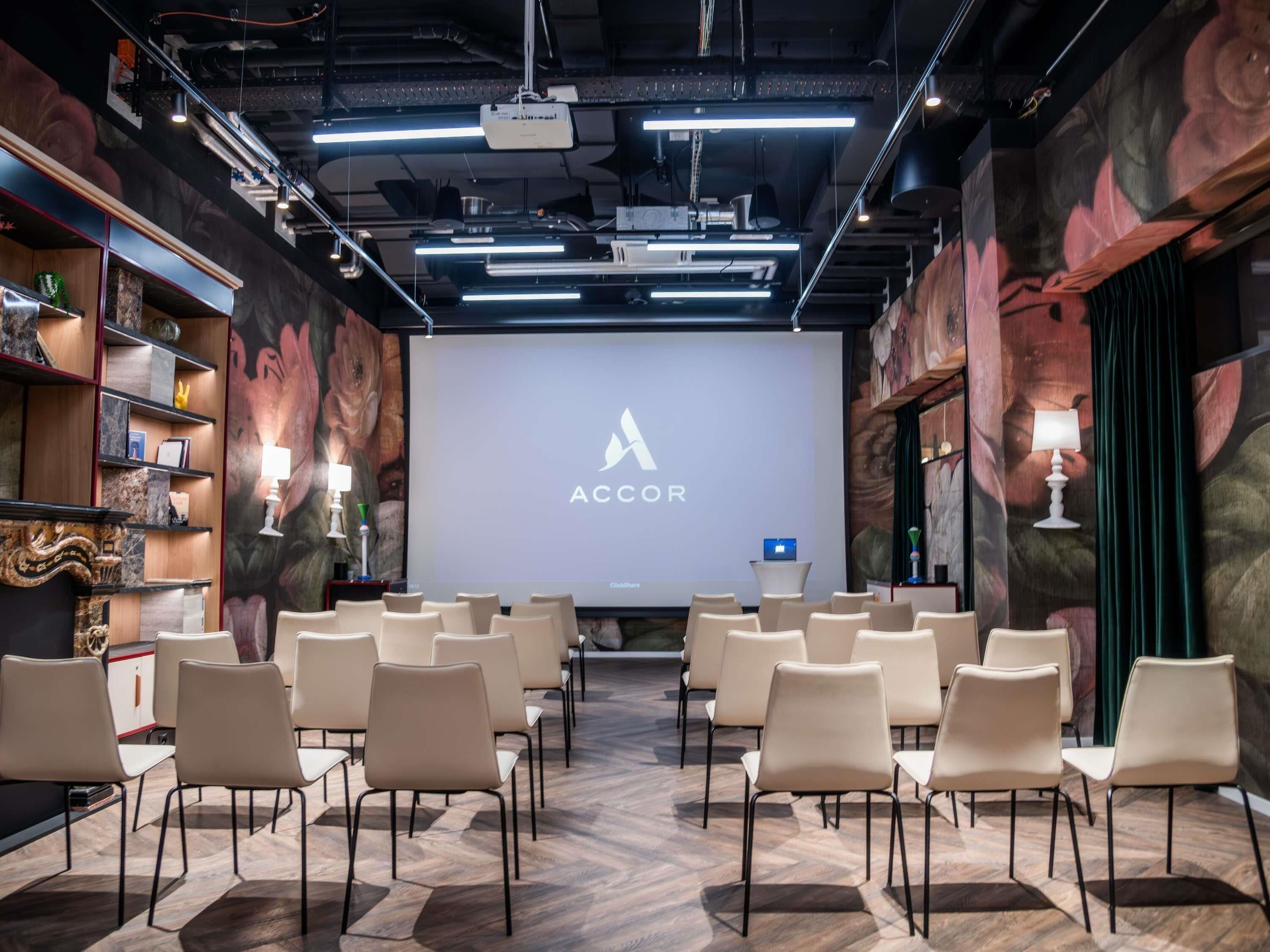 A conference room with a projector screen displaying the ACCOR logo, arranged chairs facing the screen, and a wooden floor.