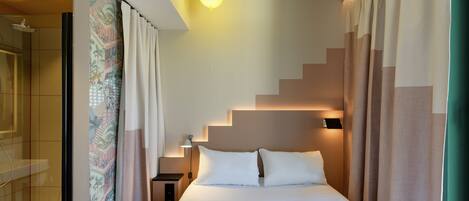 Una habitación de hotel moderna con una cama grande, una mesita de noche, una lámpara y un baño con ducha.