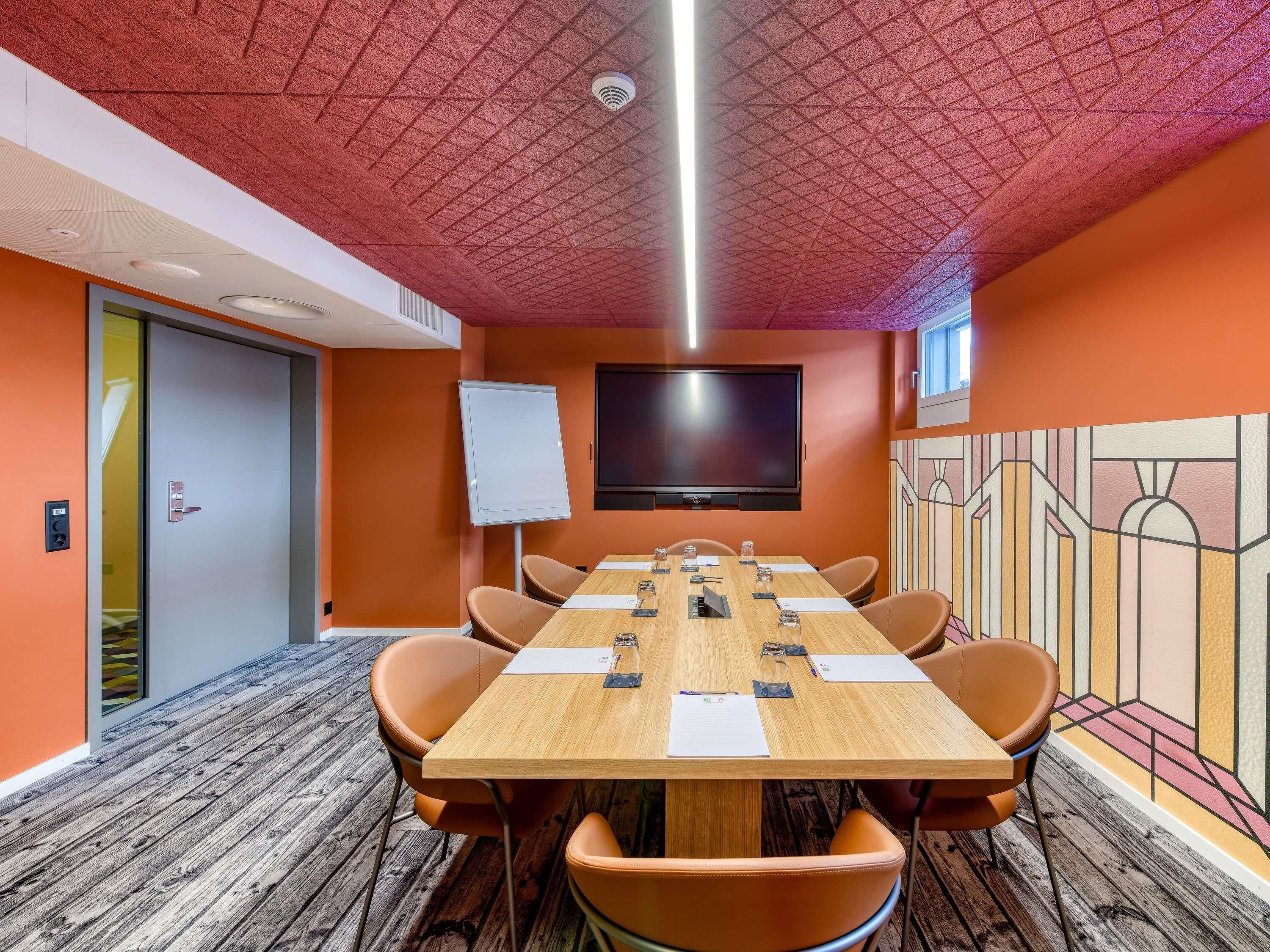 Une salle de conférence avec une longue table, des chaises, un tableau blanc et un téléviseur à écran plat fixé au mur.