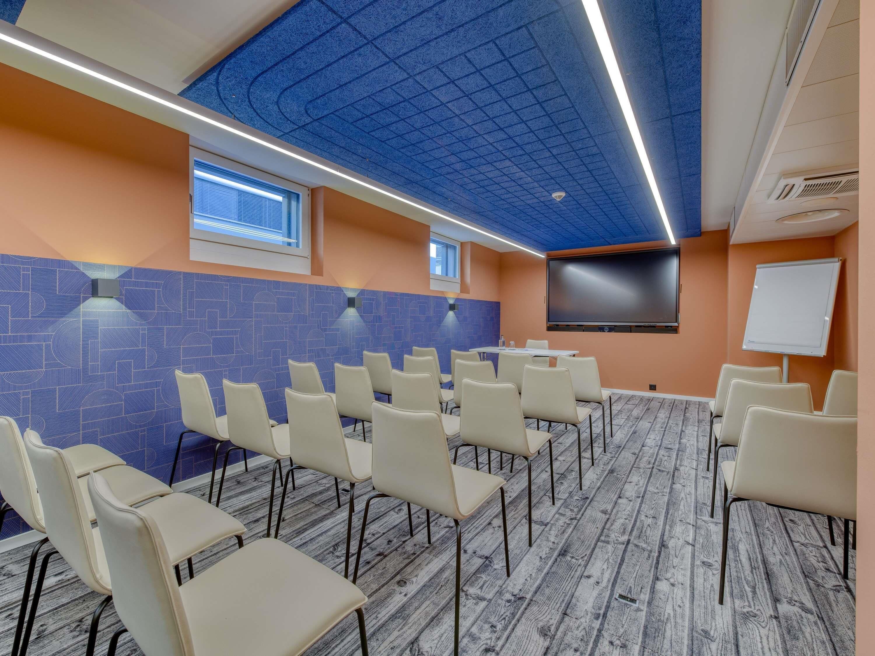 Un salón de conferencias con una pared azul estampada, una pizarra, una pantalla y filas de sillas.