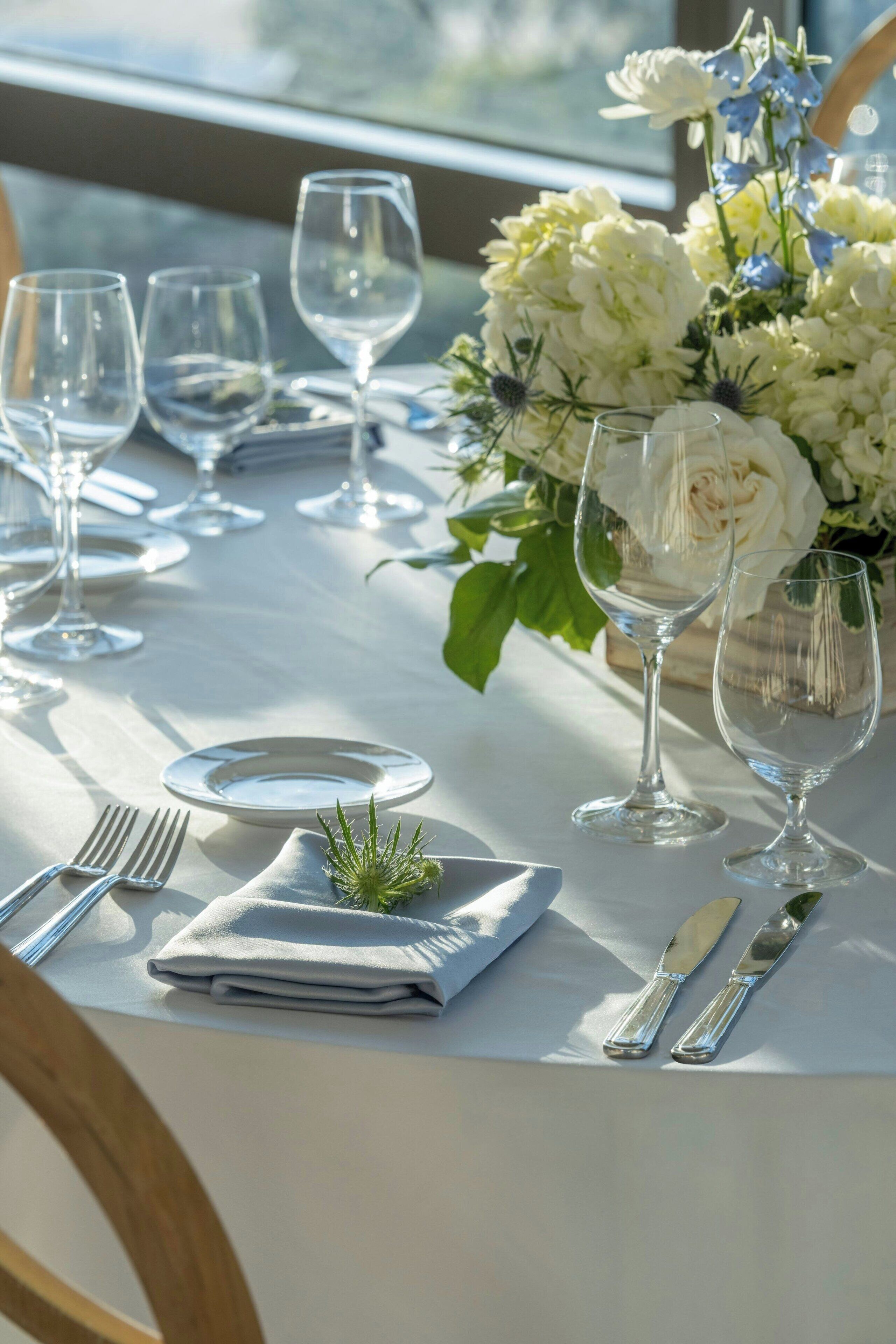 Une table est dressée pour le repas, avec de l’argenterie, des assiettes blanches et des verres à vin transparents. Un vase contenant des fleurs blanches et bleues est placé sur la table.