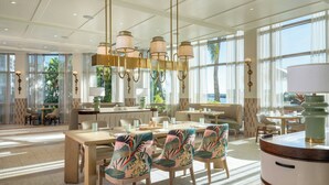 A spacious hotel lobby with a large dining area, modern chandeliers, and large windows offering views of the outdoors.