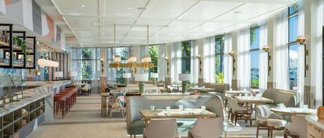 Um hall de hotel espaçoso, com teto alto, amplas janelas e diversas opções de assentos, incluindo cabines e mesas.
