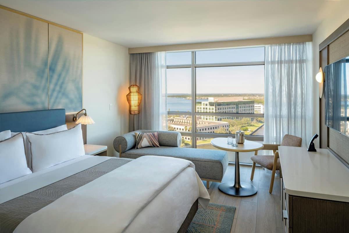A hotel room with a large bed, a sofa, a dining table, and a view of the cityscape.