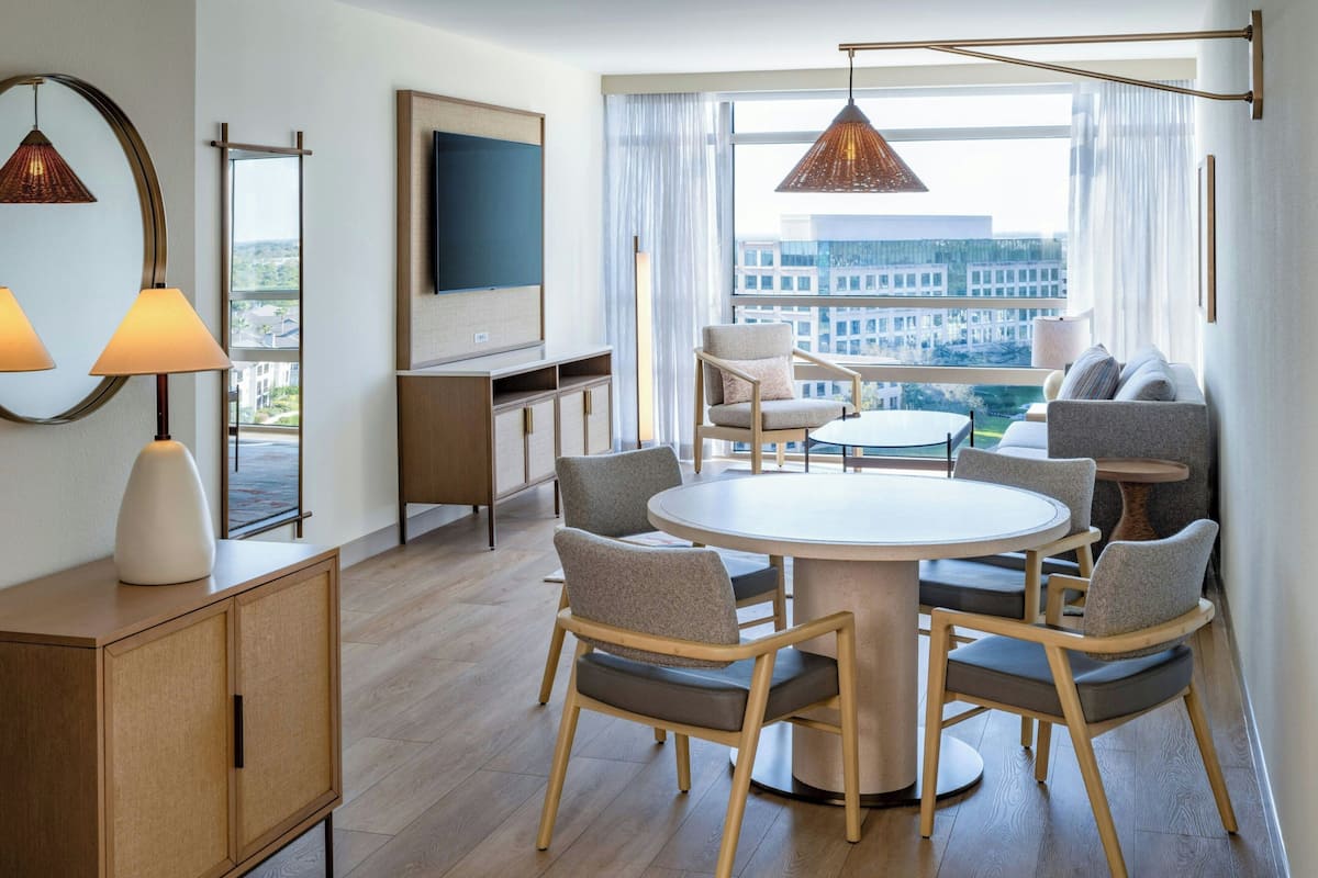 A modern hotel room with a round dining table, four chairs, a TV, a large window with curtains, and a city view.