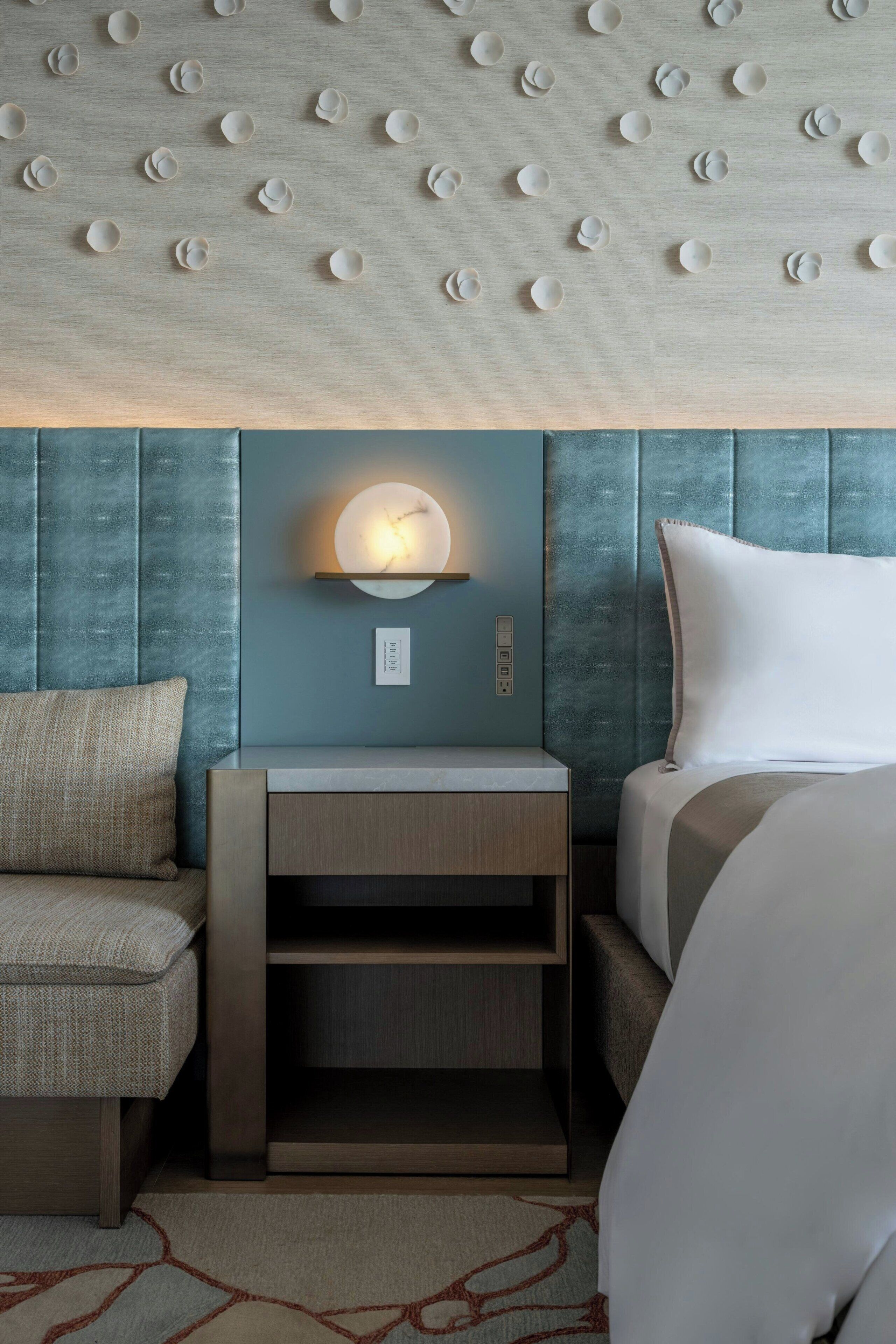 A hotel room with a bed, a nightstand, a wall-mounted light, and a textured wall panel.