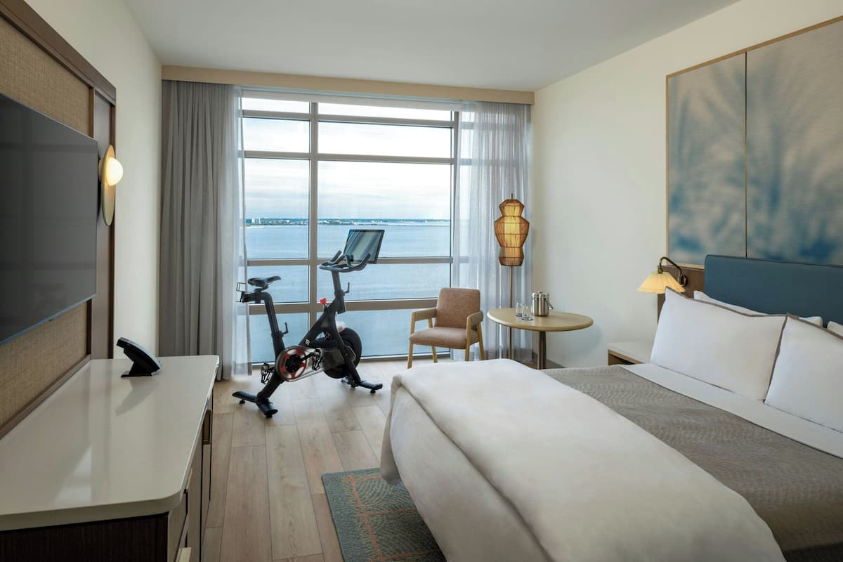 A hotel room with a bed, desk, chair, exercise bike, and a view of the sea.