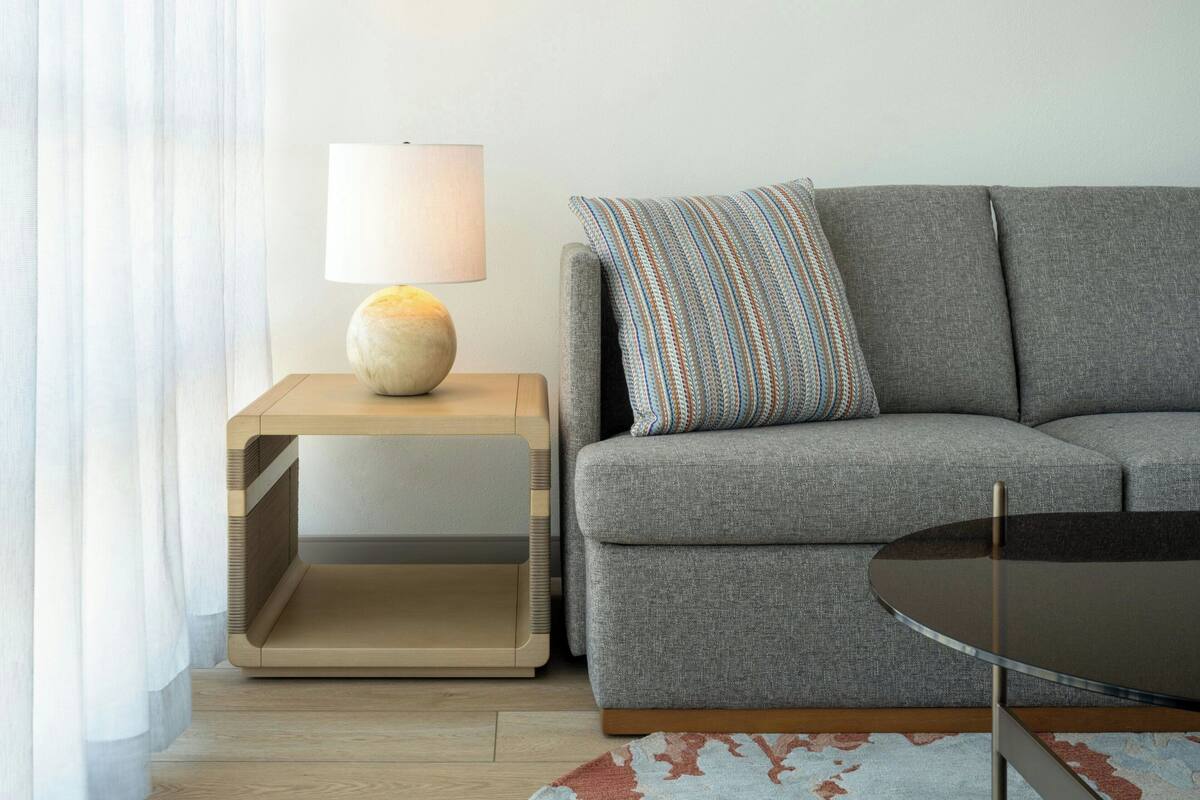 A modern living room with a grey sofa, a round glass coffee table, a side table with a lamp, and a patterned throw pillow.