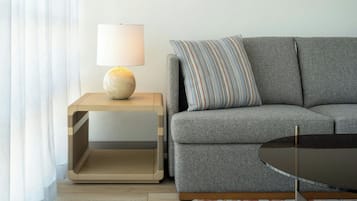 A modern living room with a grey sofa, a round glass coffee table, a side table with a lamp, and a patterned throw pillow.