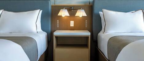 Una habitación de hotel con dos camas, una mesita de noche con lámpara y un estante pequeño.