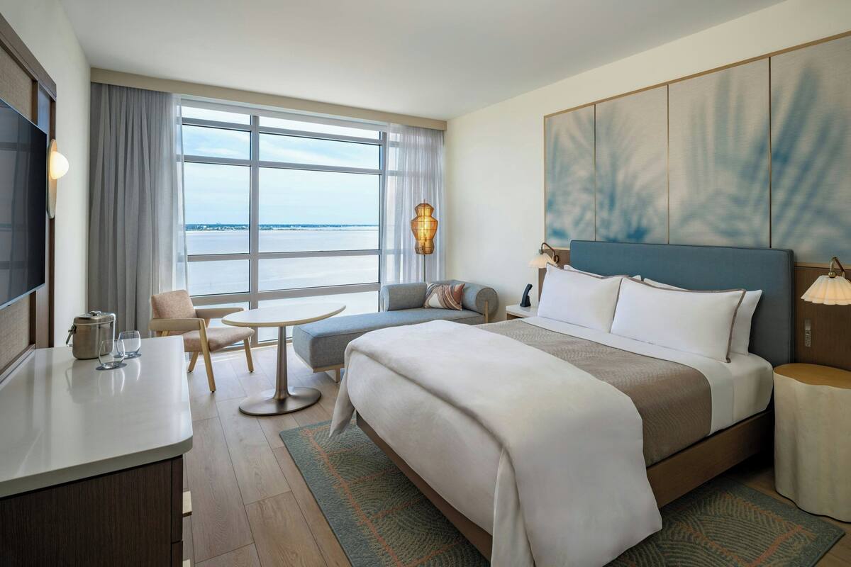 A hotel room with a large bed, a desk with a TV, a seating area with a table and chairs, and a view of the water through large windows.