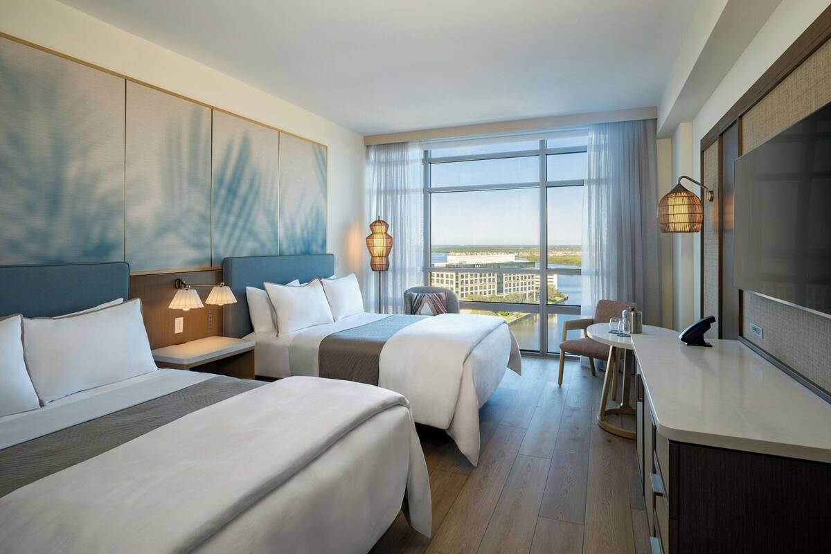 A hotel room with two beds, a desk, a chair, and a large window offering a city view.