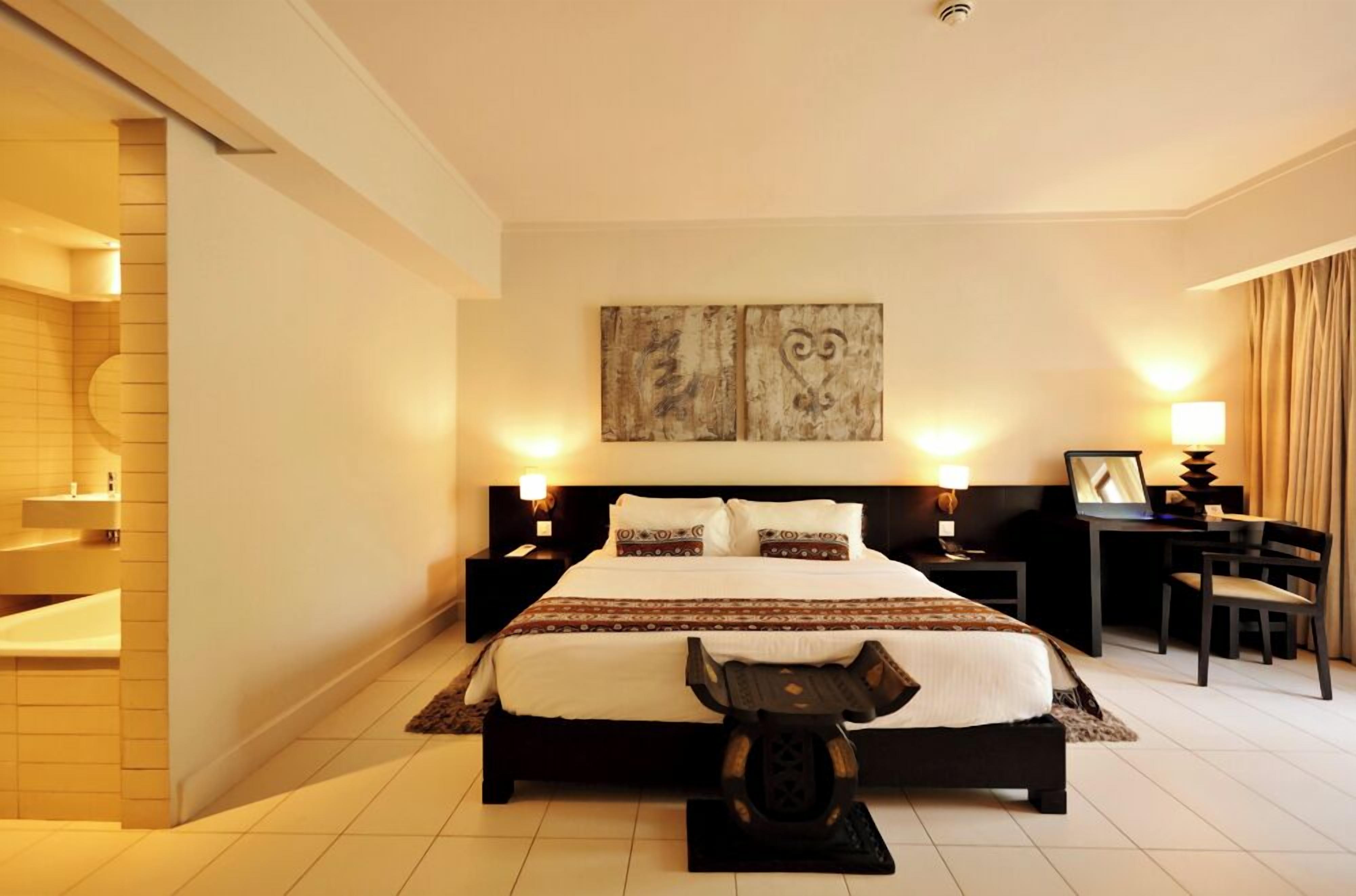 A hotel room with a large bed, bedside tables, a desk, a chair, and artwork on the wall.