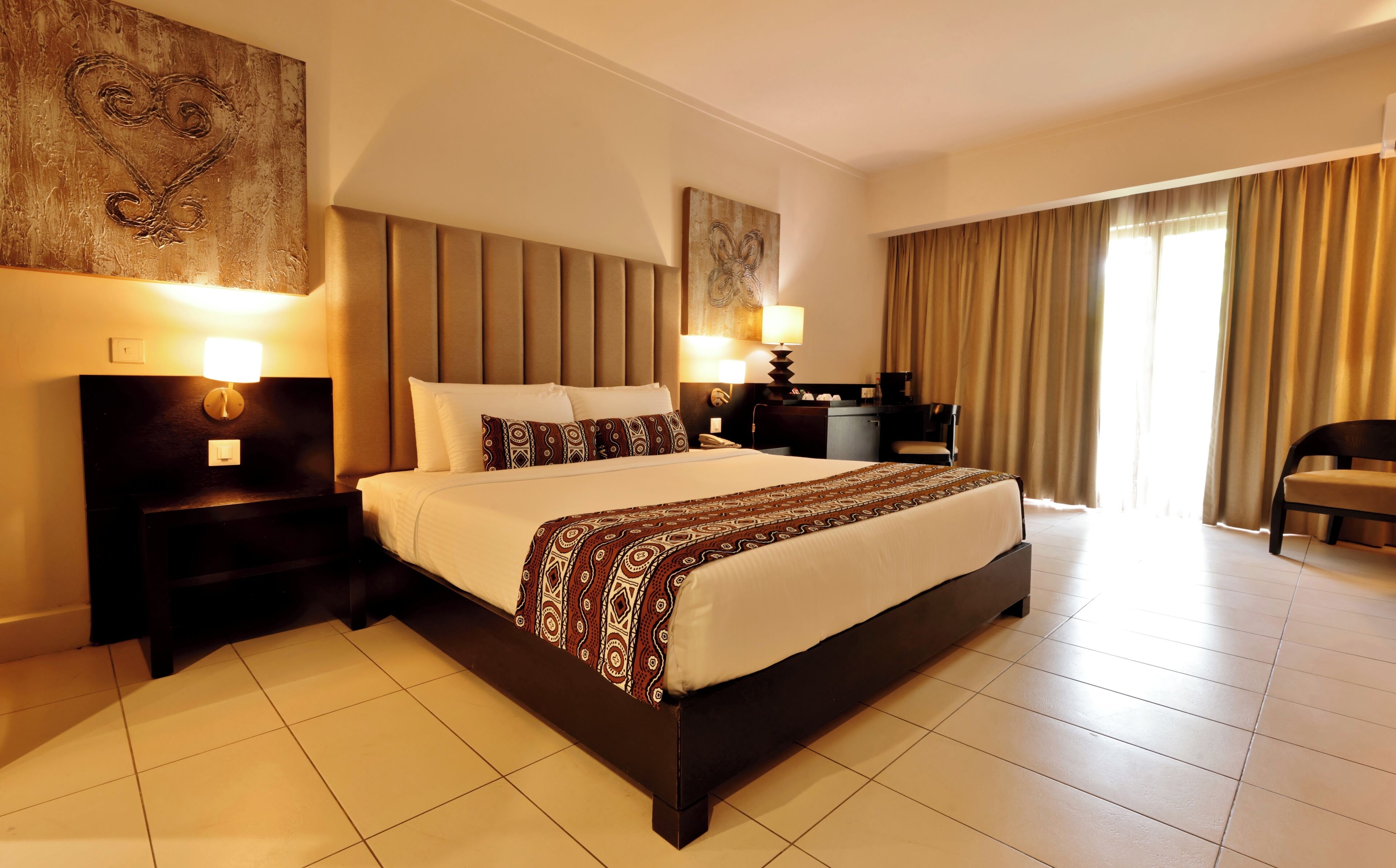 A hotel room with a large bed, two bedside tables with lamps, a desk with a chair, and a large window with curtains.