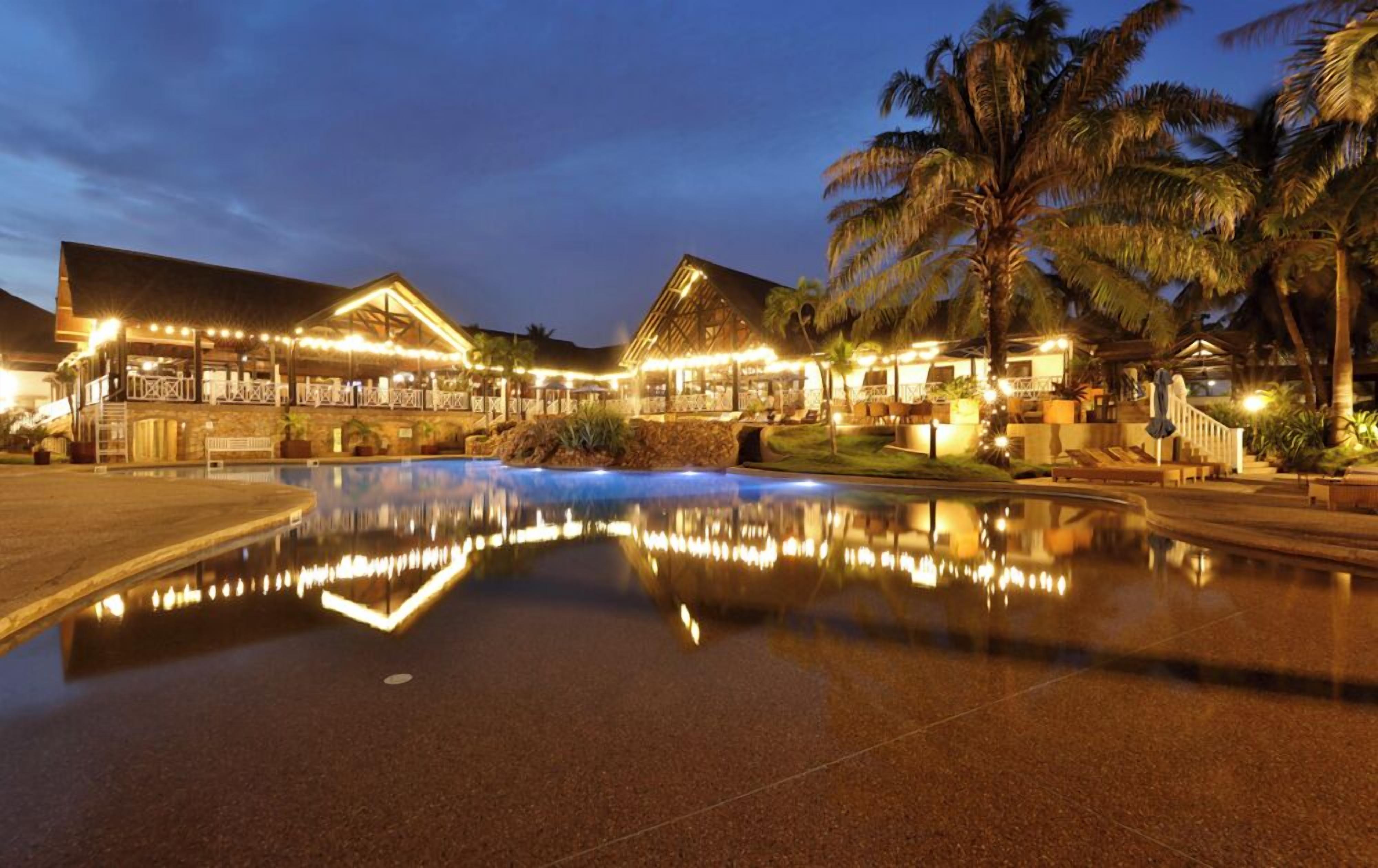 A resort with a large pool reflecting the buildings and palm trees, a covered outdoor area with seating, and a well-lit entrance.