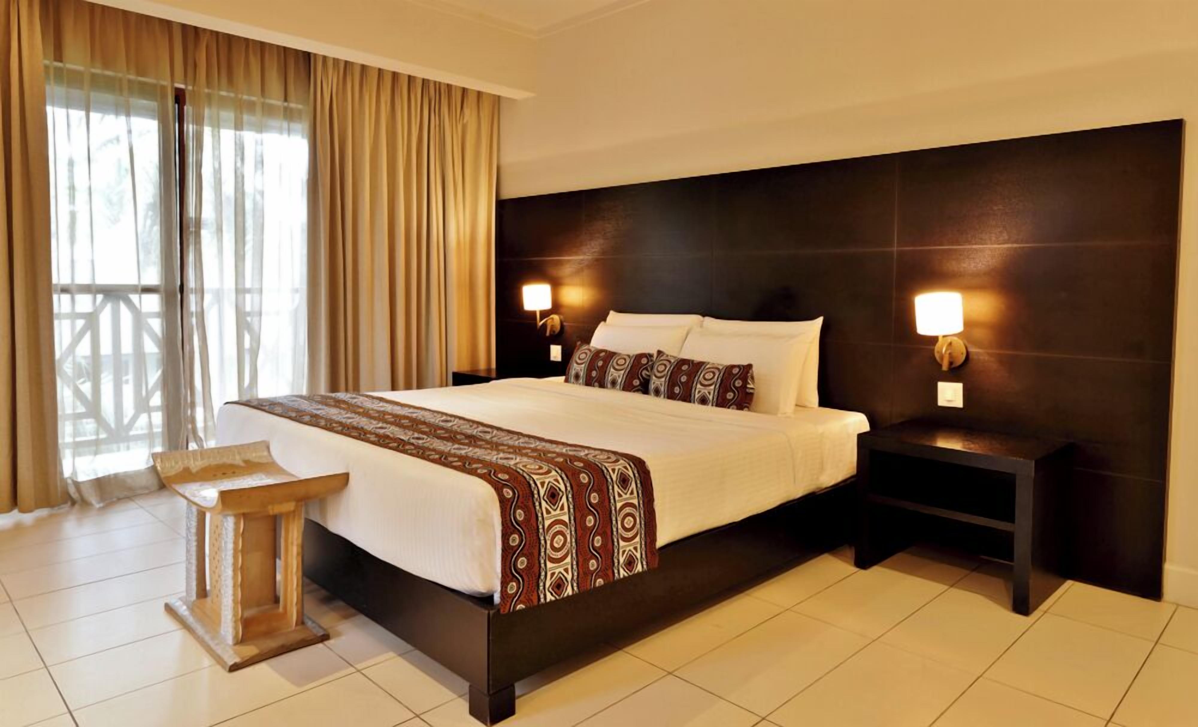 A hotel room with a large bed, two bedside tables with lamps, a small wooden side table, and a view of the outdoors through a large window with curtains.