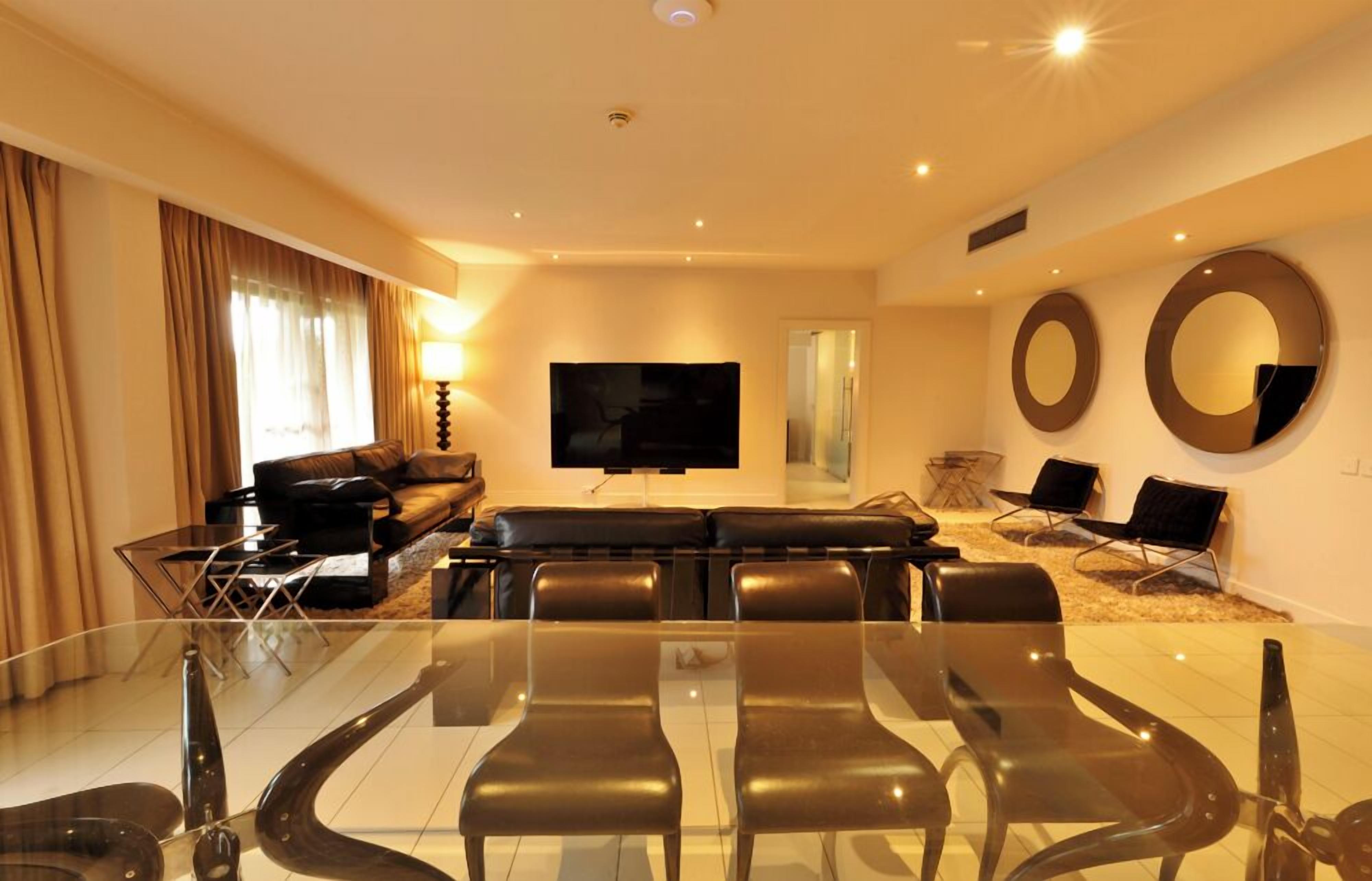 A modern living room with a glass dining table, black leather sofa, and a flat-screen TV.