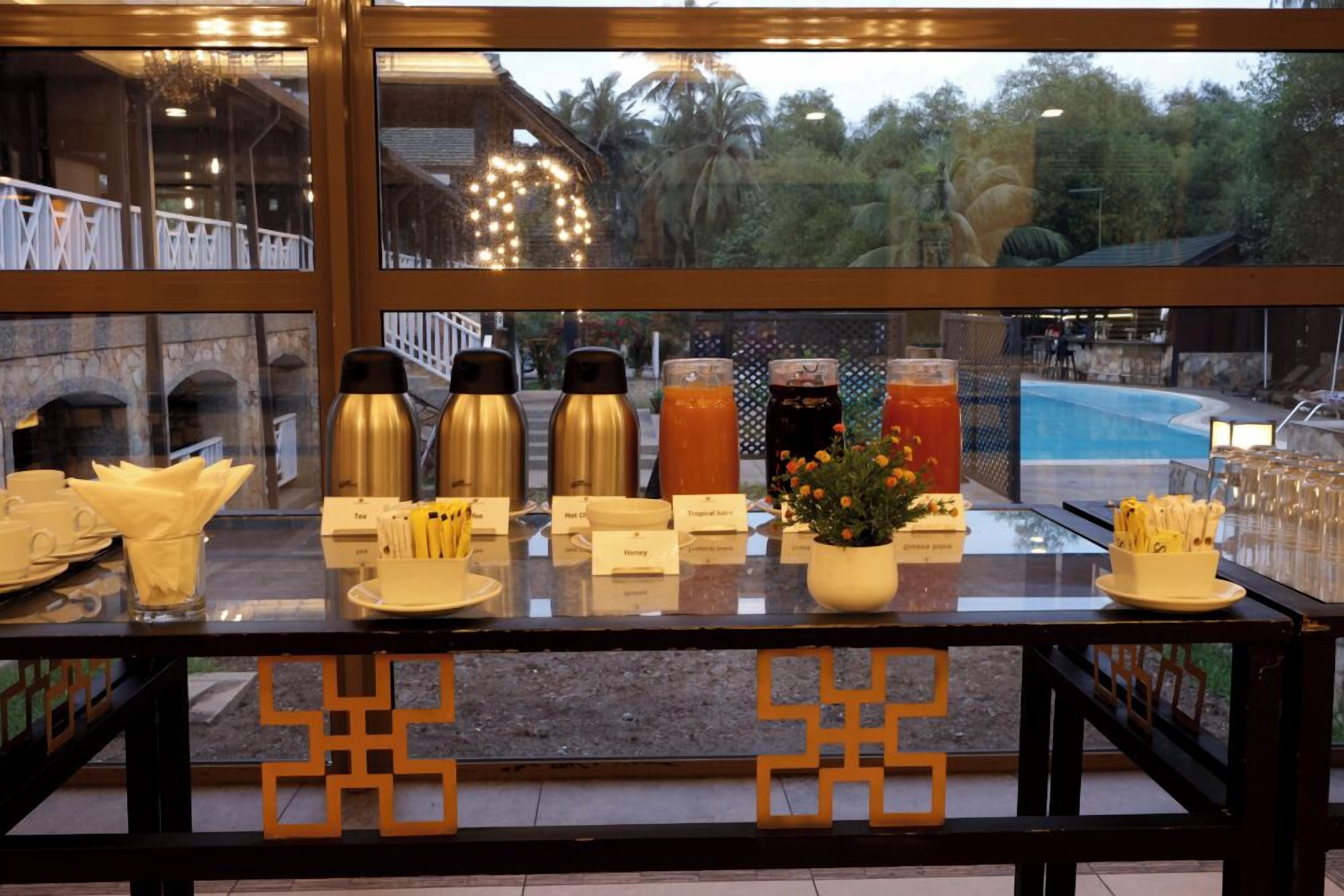 A buffet setup with various food and drink items, including hot beverages and juices, arranged on a glass table with a view of a pool and greenery outside.