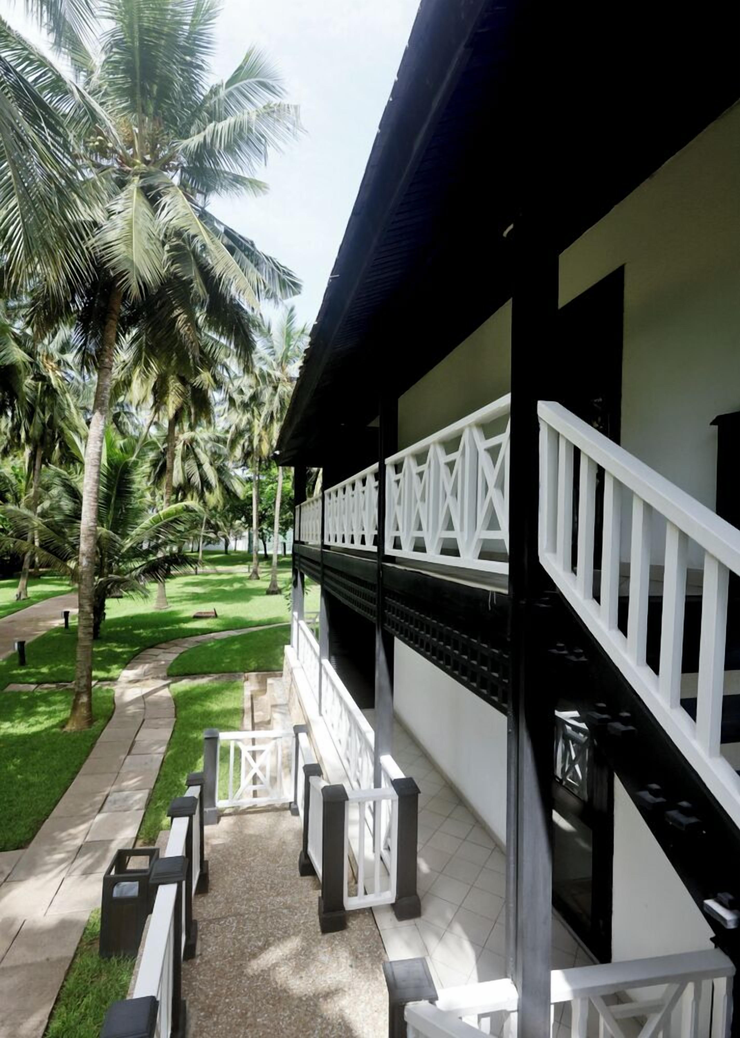 A covered outdoor area with a white railing, palm trees, and a pathway leading to a building entrance.