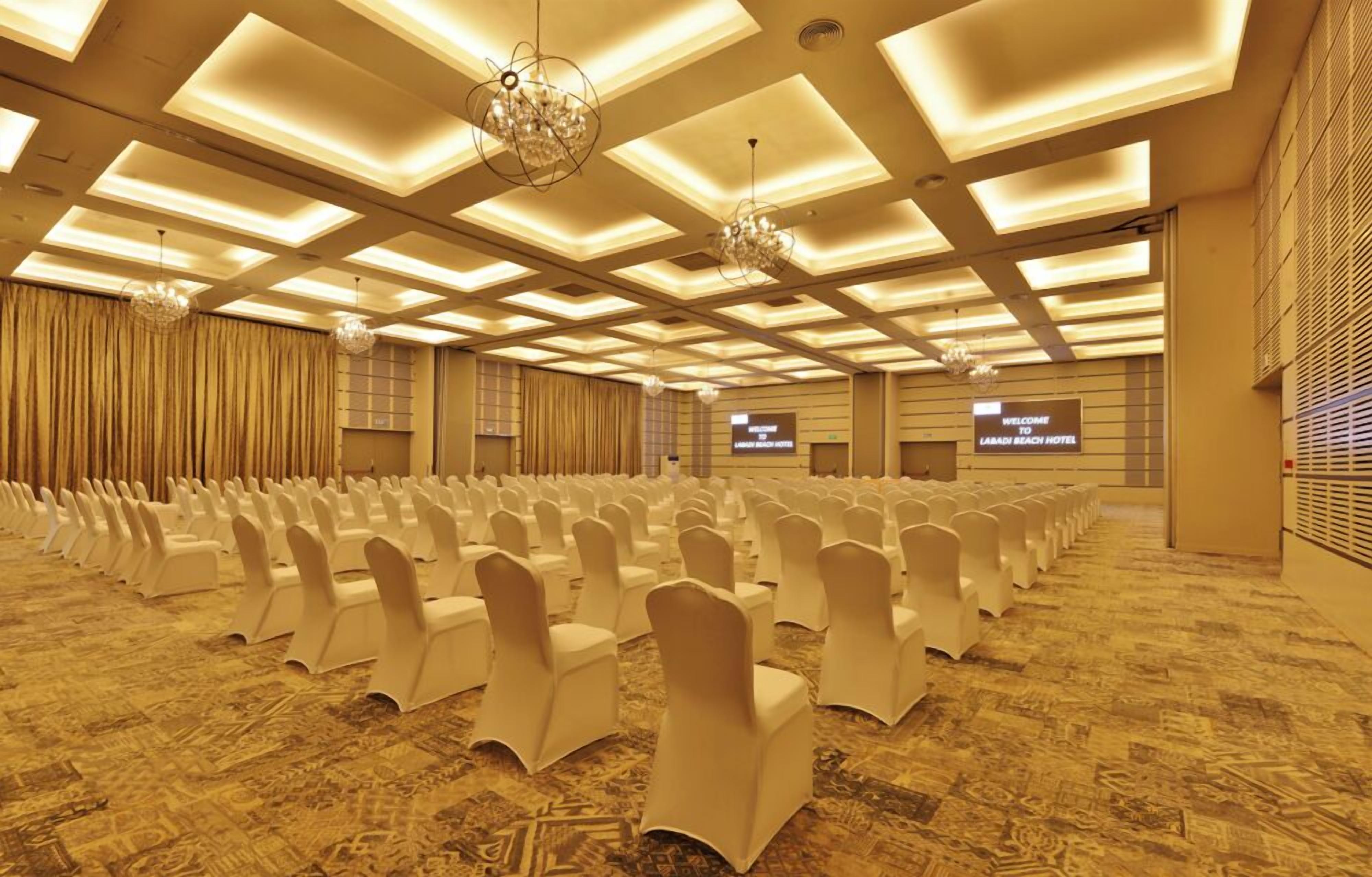 A large conference room with rows of white chairs, a patterned carpet, and a ceiling with recessed lighting and chandeliers.